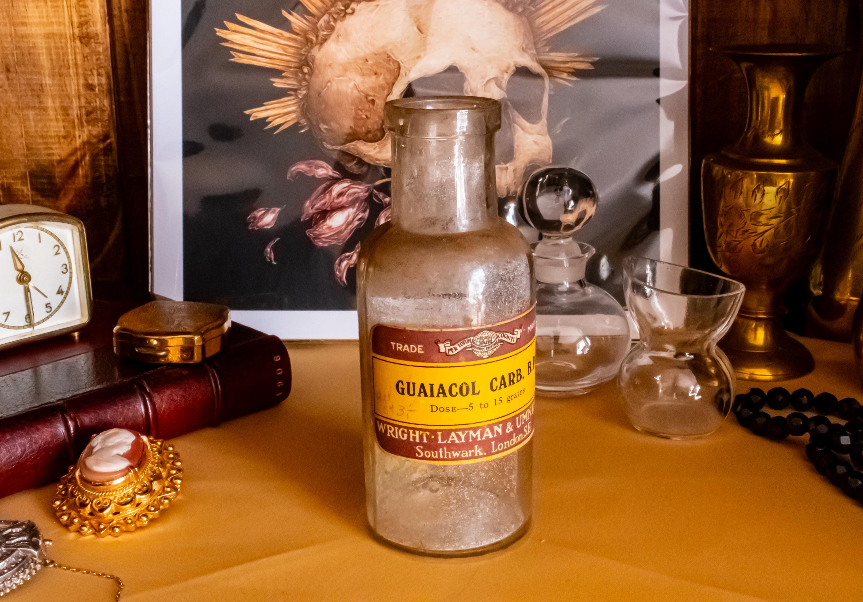 Antique clear glass apothecary bottle with a yellow and red paper label reading "Guaiacol Carb" from London, sitting next to a vintage glass stopper and an anatomical skull print.