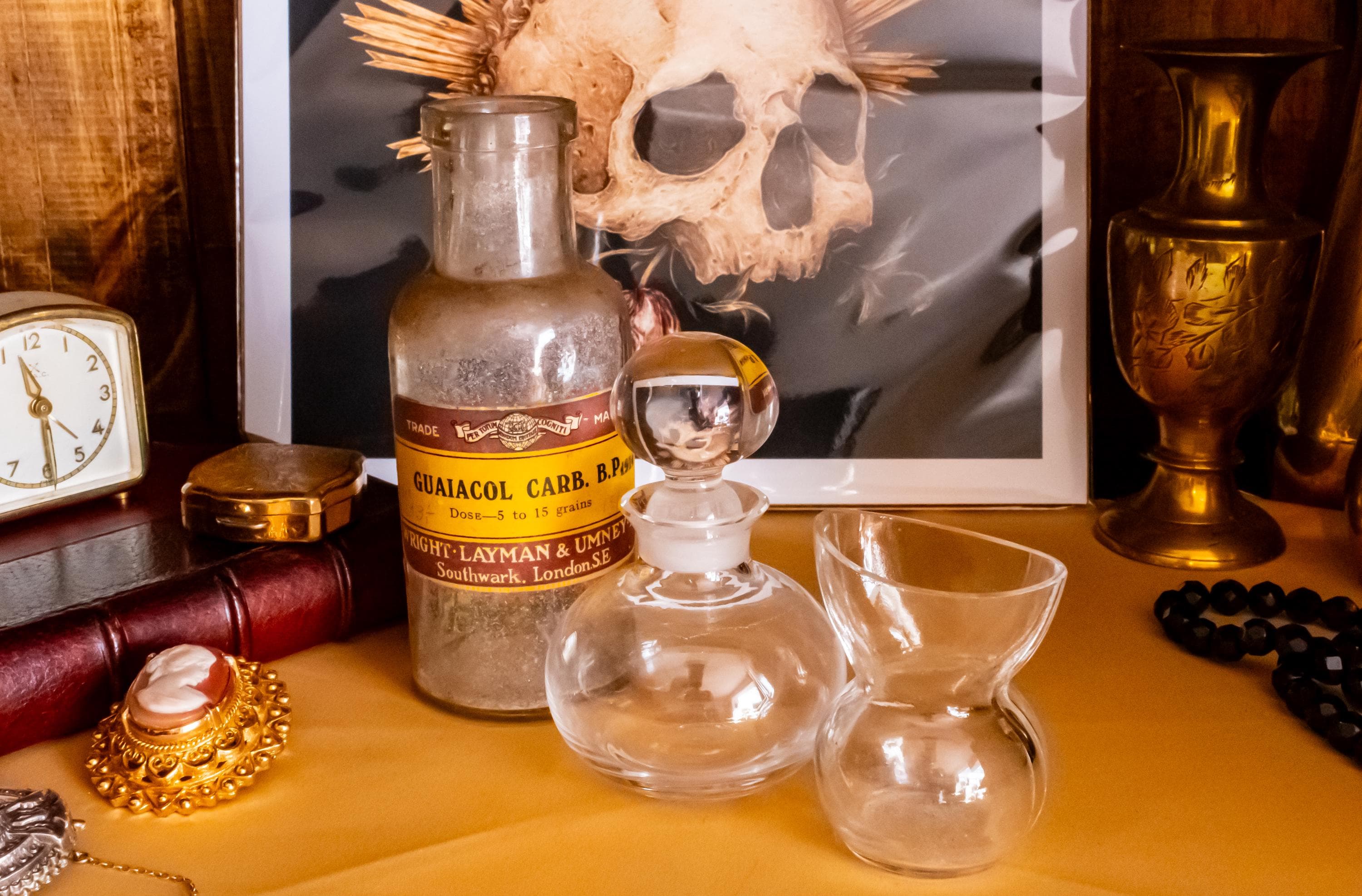 Antique clear glass apothecary bottle with a yellow and red paper label reading "Guaiacol Carb" from London, sitting next to a vintage glass stopper and an anatomical skull print.