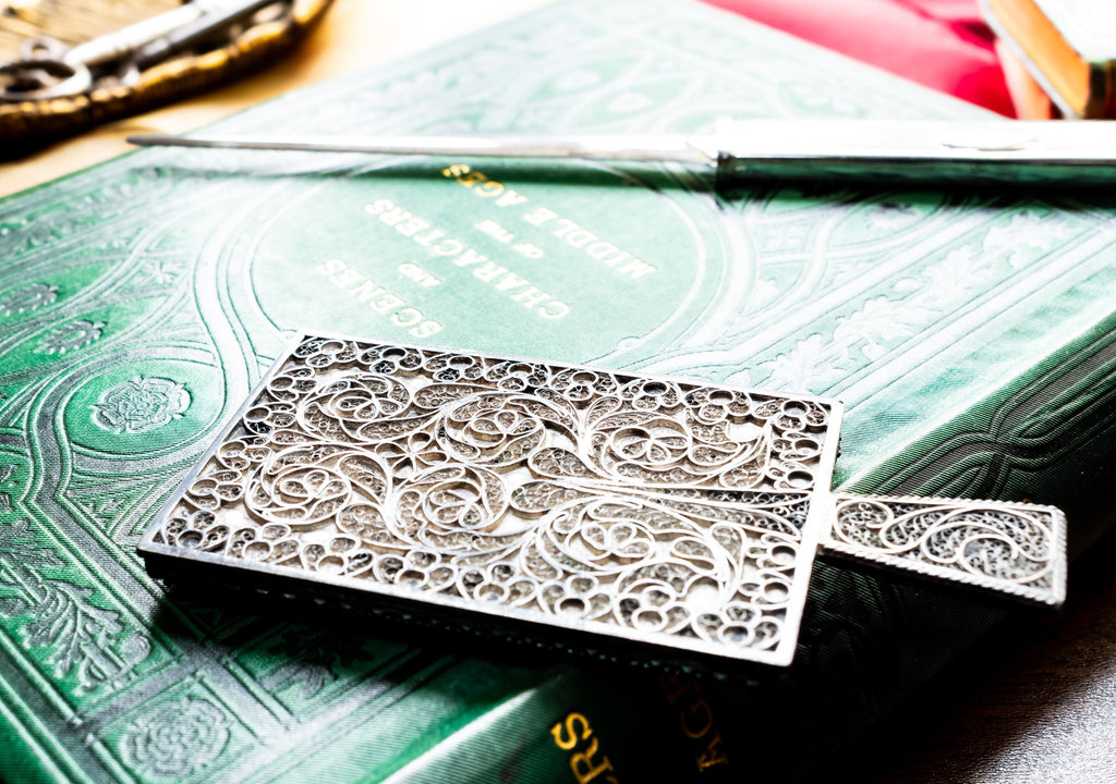 Antique rectangular silver hand mirror featuring intricate swirling filigree wirework on the back and a beveled glass front, resting on a green vintage book.