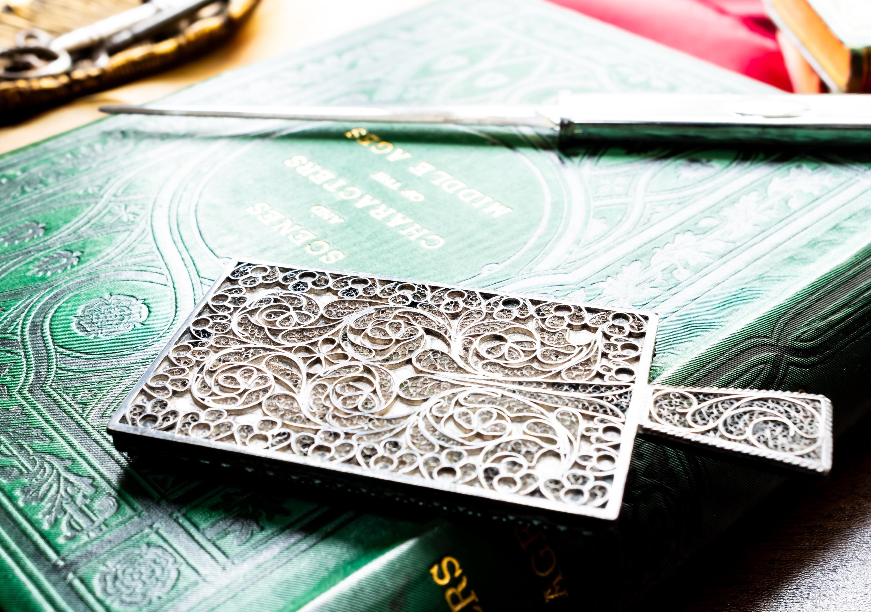 Antique rectangular silver hand mirror featuring intricate swirling filigree wirework on the back and a beveled glass front, resting on a green vintage book.