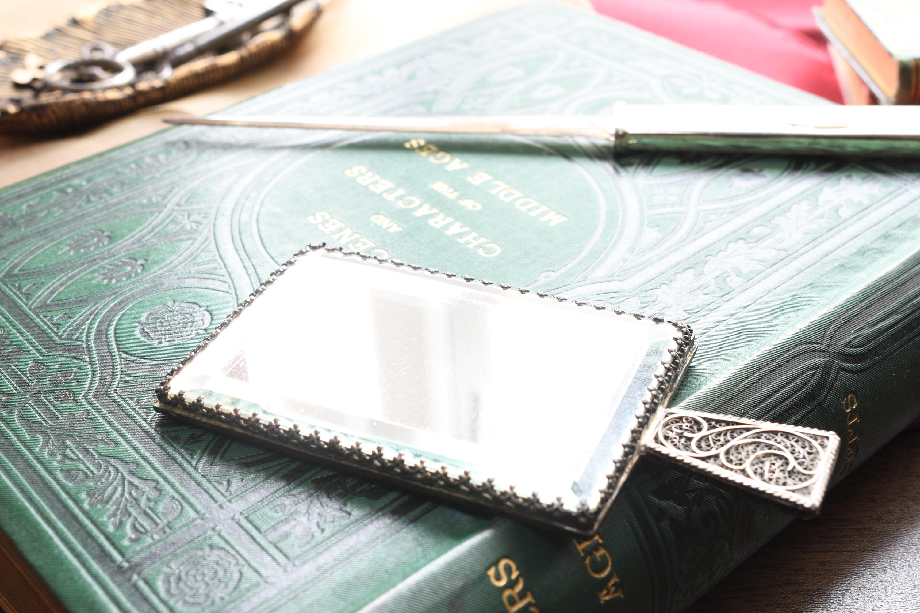 Antique rectangular silver hand mirror featuring intricate swirling filigree wirework on the back and a beveled glass front, resting on a green vintage book.