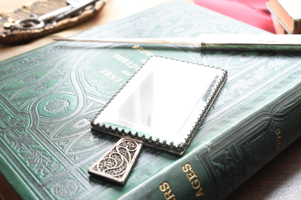 Antique rectangular silver hand mirror featuring intricate swirling filigree wirework on the back and a beveled glass front, resting on a green vintage book.
