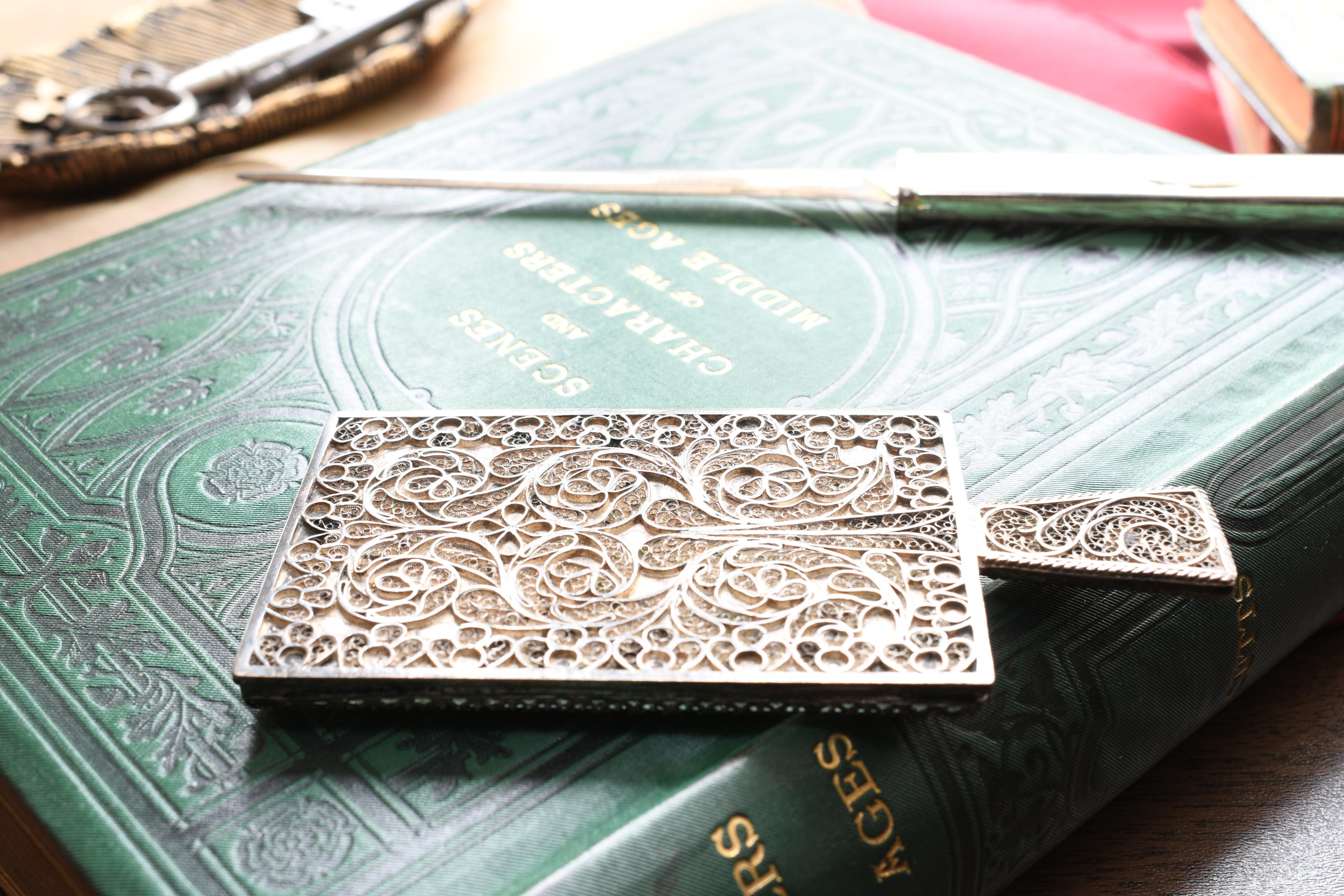 Antique rectangular silver hand mirror featuring intricate swirling filigree wirework on the back and a beveled glass front, resting on a green vintage book.