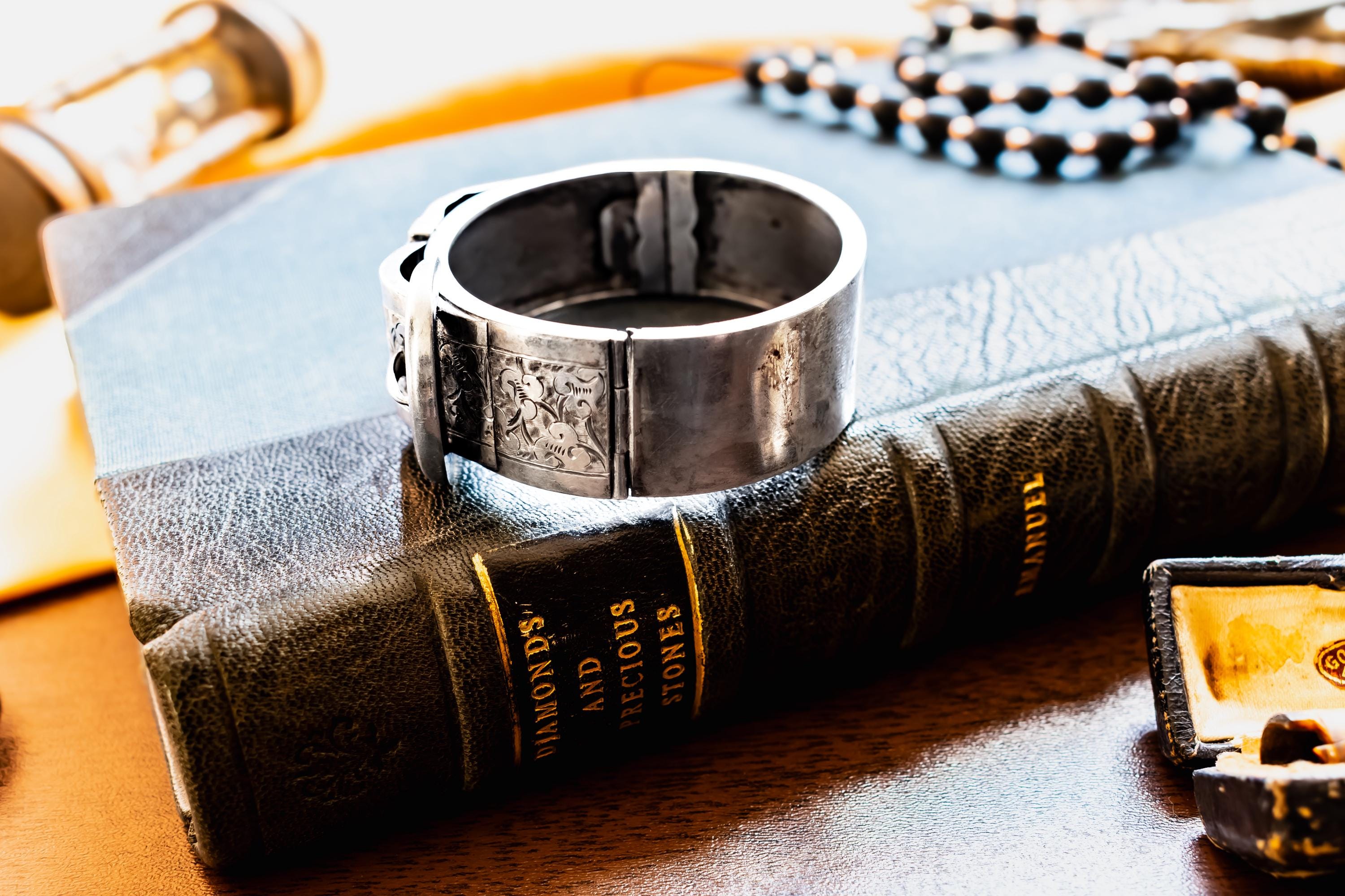 Chunky antique silver Victorian hinged bangle bracelet shaped like a buckled belt with floral engravings, resting on a dark vintage book titled Diamonds and Precious Stones.