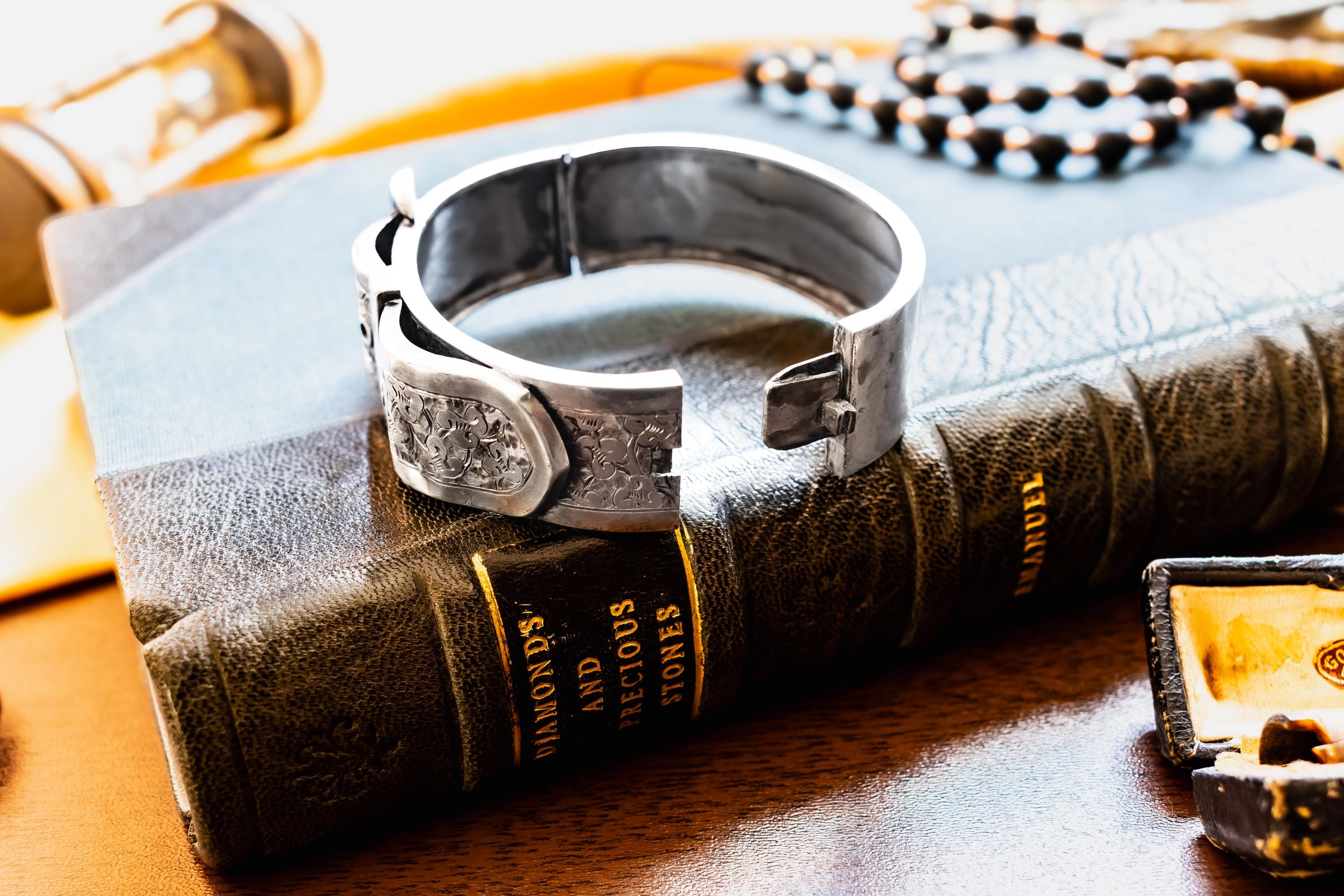 Chunky antique silver Victorian hinged bangle bracelet shaped like a buckled belt with floral engravings, resting on a dark vintage book titled Diamonds and Precious Stones.