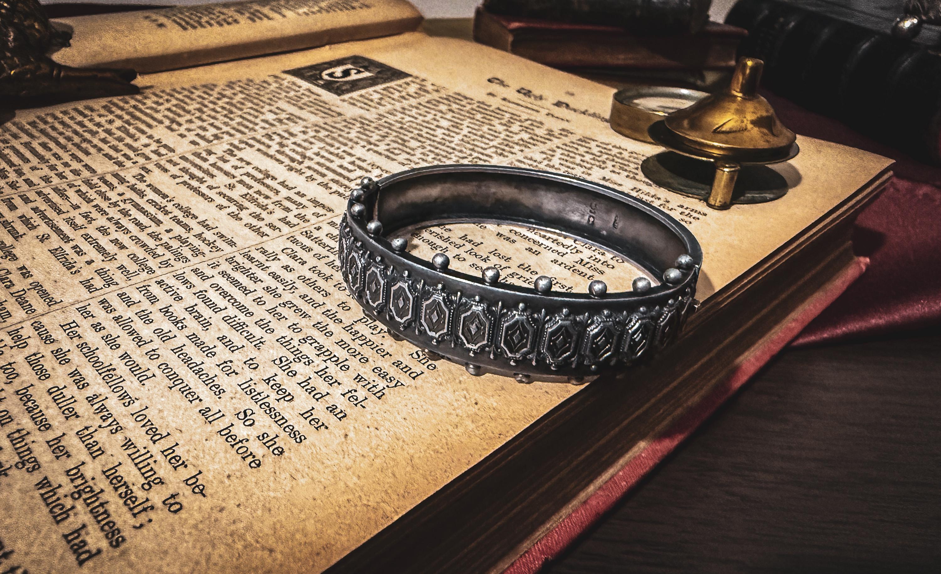 An antique 1883 heavy sterling silver cuff bracelet by Robert James Dick featuring intricate geometric and beadwork detailing, resting on the text of an open vintage book.