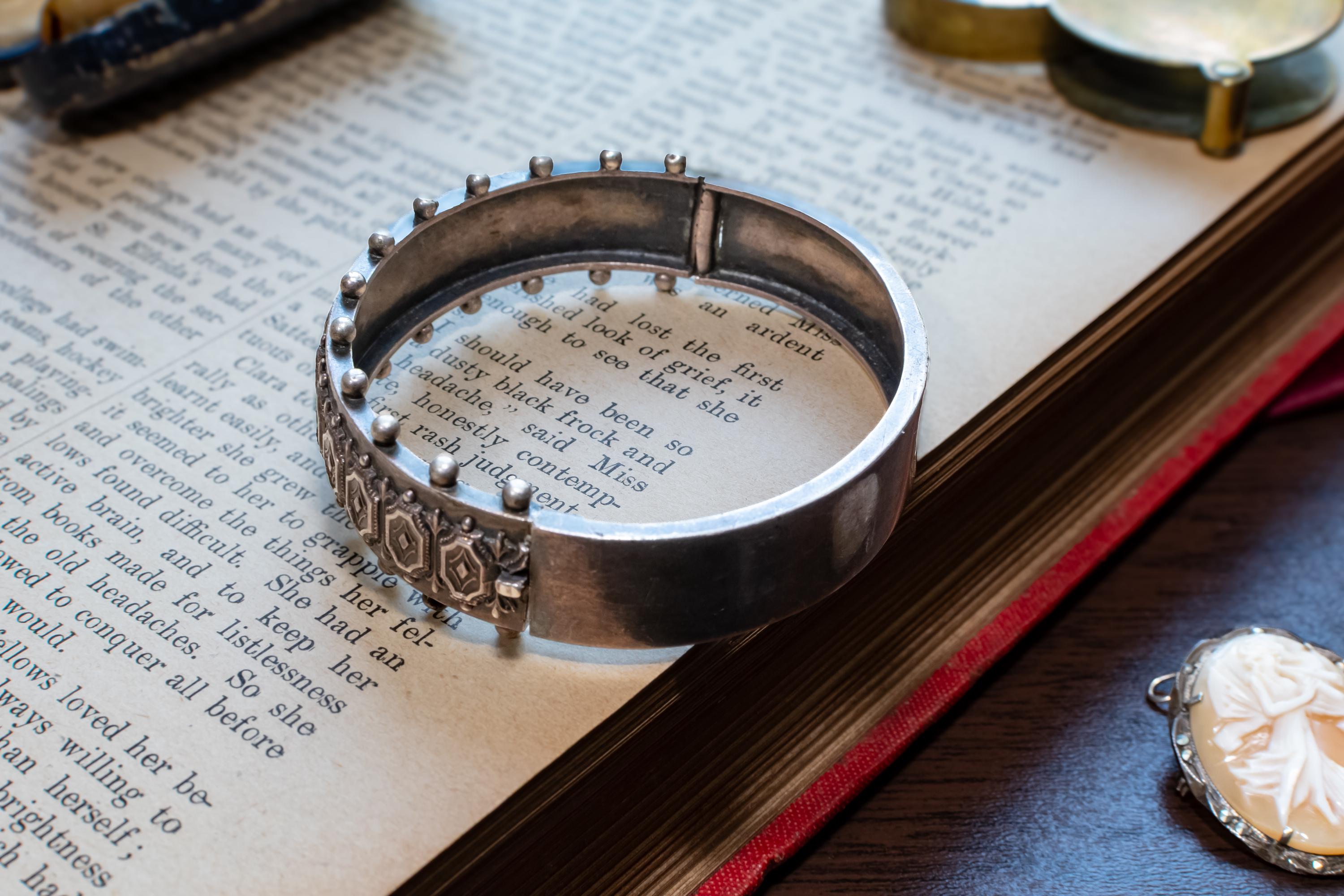 An antique 1883 heavy sterling silver cuff bracelet by Robert James Dick featuring intricate geometric and beadwork detailing, resting on the text of an open vintage book.