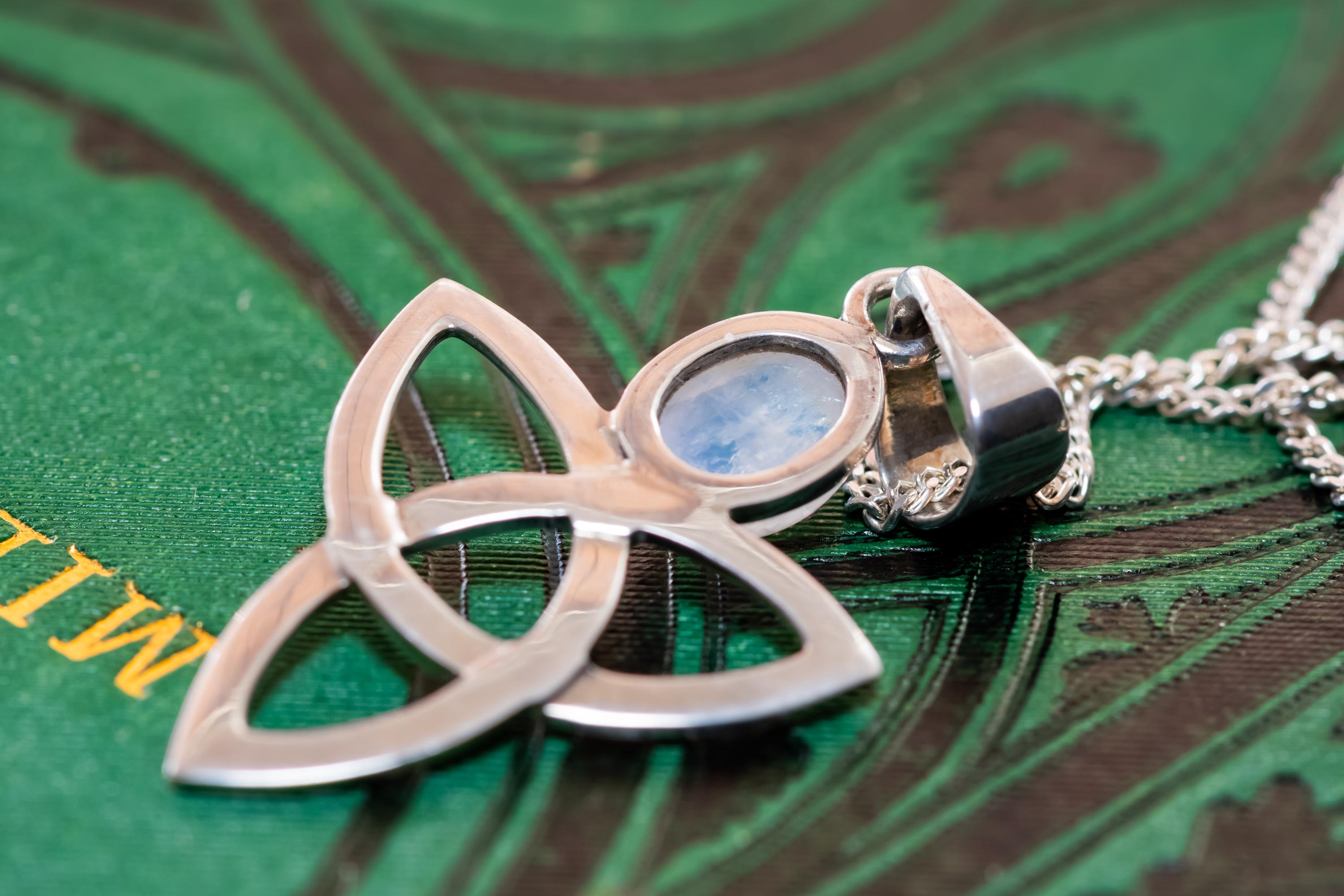Close-up of a vintage 925 sterling silver pendant necklace featuring a Celtic triquetra knot and a glowing oval moonstone cabochon, resting elegantly on a dark green book cover with gold lettering