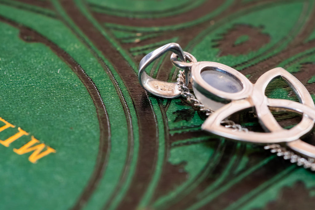 Close-up of a vintage 925 sterling silver pendant necklace featuring a Celtic triquetra knot and a glowing oval moonstone cabochon, resting elegantly on a dark green book cover with gold lettering