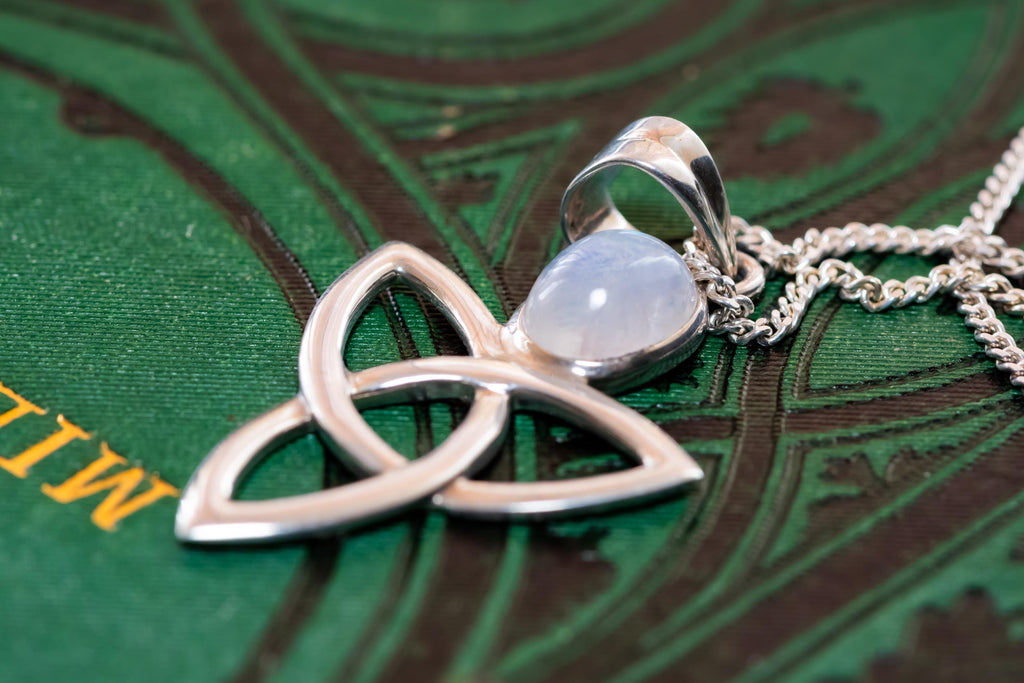 Close-up of a vintage 925 sterling silver pendant necklace featuring a Celtic triquetra knot and a glowing oval moonstone cabochon, resting elegantly on a dark green book cover with gold lettering