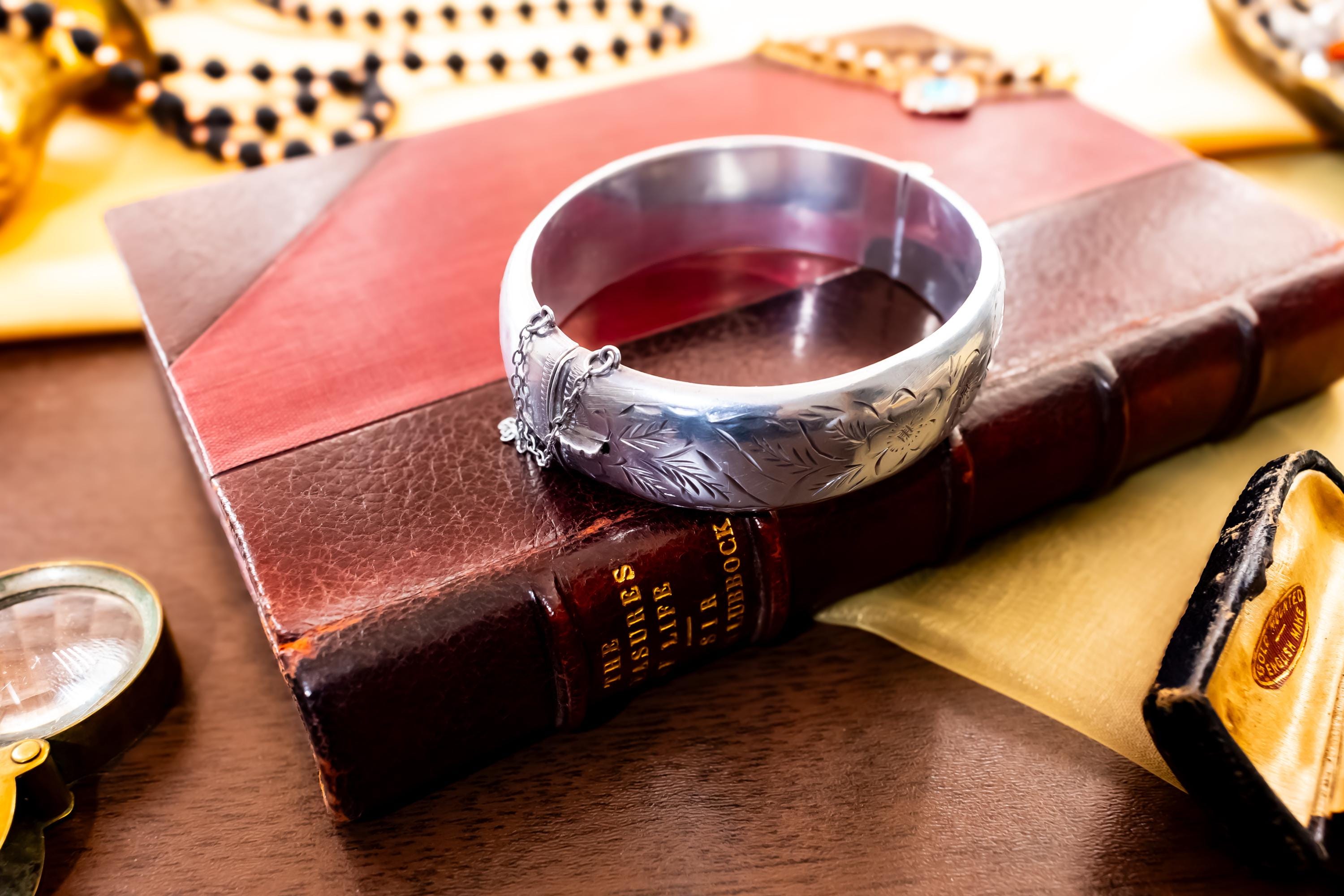 A wide vintage 1969 Birmingham sterling silver hinged bangle with intricate botanical engravings and a safety chain, resting beautifully on a vintage leather-bound book.
