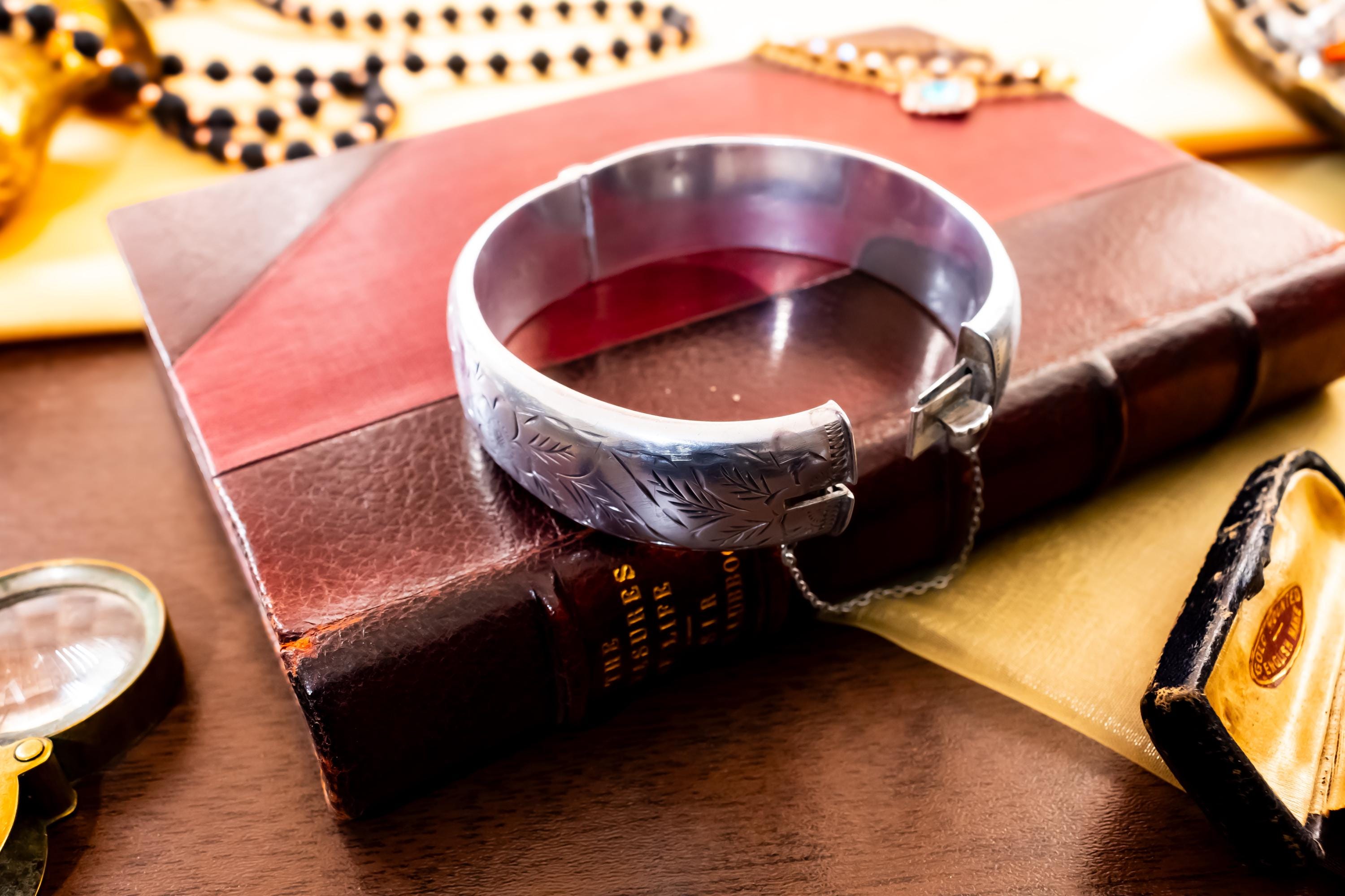 Close-up of a wide, vintage 1969 Birmingham sterling silver hinged bangle with intricate botanical engravings and a safety chain, resting beautifully on a vintage leather-bound book.