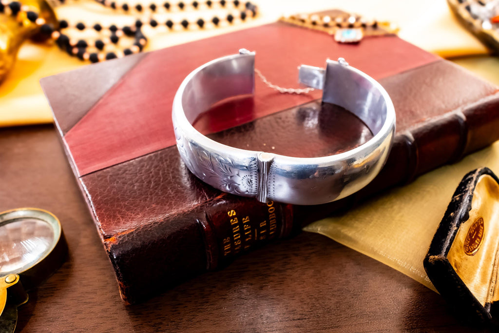 Close-up of a wide, vintage 1969 Birmingham sterling silver hinged bangle with intricate botanical engravings and a safety chain, resting beautifully on a vintage leather-bound book.