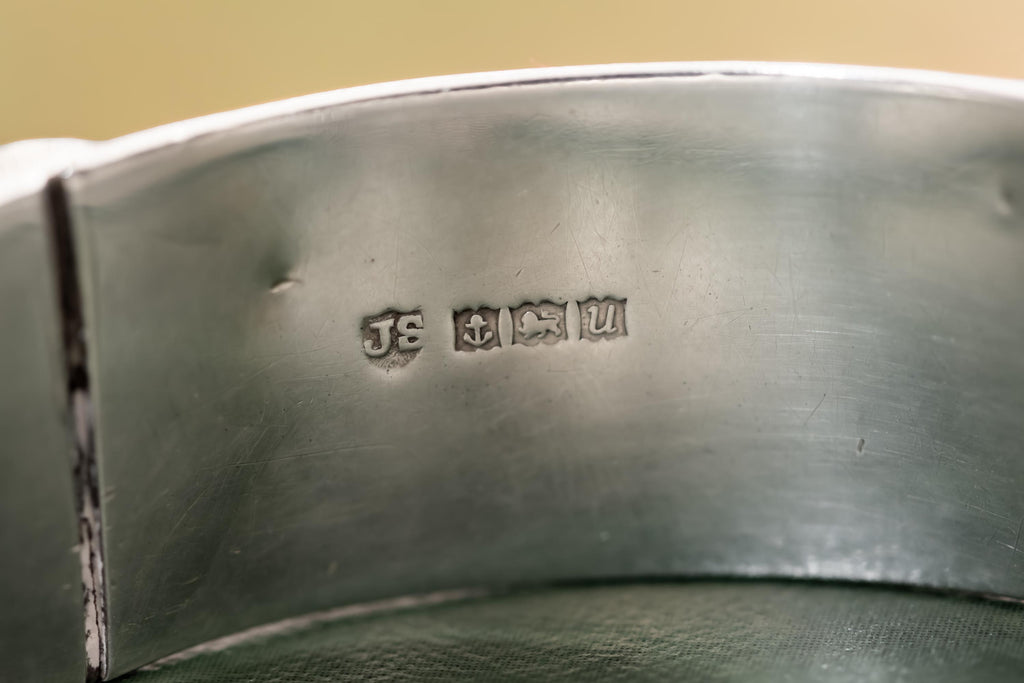 Close-up of a wide, vintage 1969 Birmingham sterling silver hinged bangle with intricate botanical engravings and a safety chain, resting beautifully on a vintage leather-bound book.