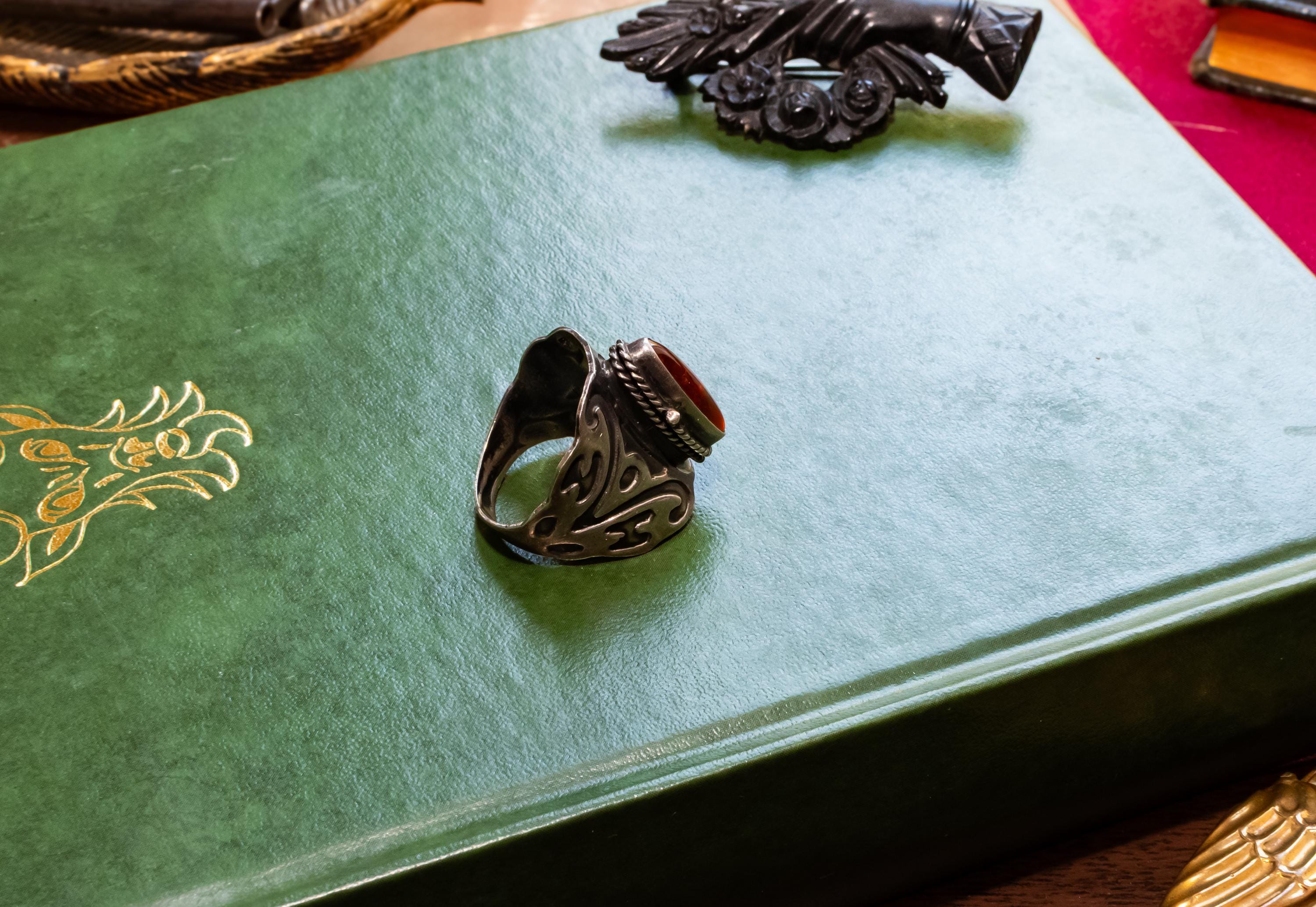 Close-up of a heavy vintage sterling silver ring featuring a dark amber cabochon top. The amber stone is hinged and slightly open to reveal a secret hidden poison locket compartment inside, set against a dark academia aesthetic background.