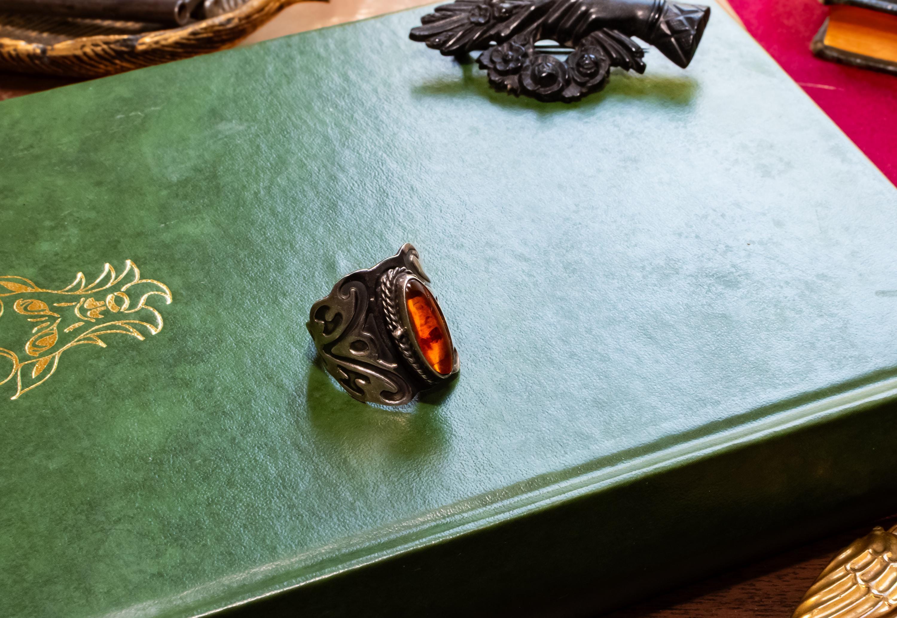 Close-up of a heavy vintage sterling silver ring featuring a dark amber cabochon top. The amber stone is hinged and slightly open to reveal a secret hidden poison locket compartment inside, set against a dark academia aesthetic background.