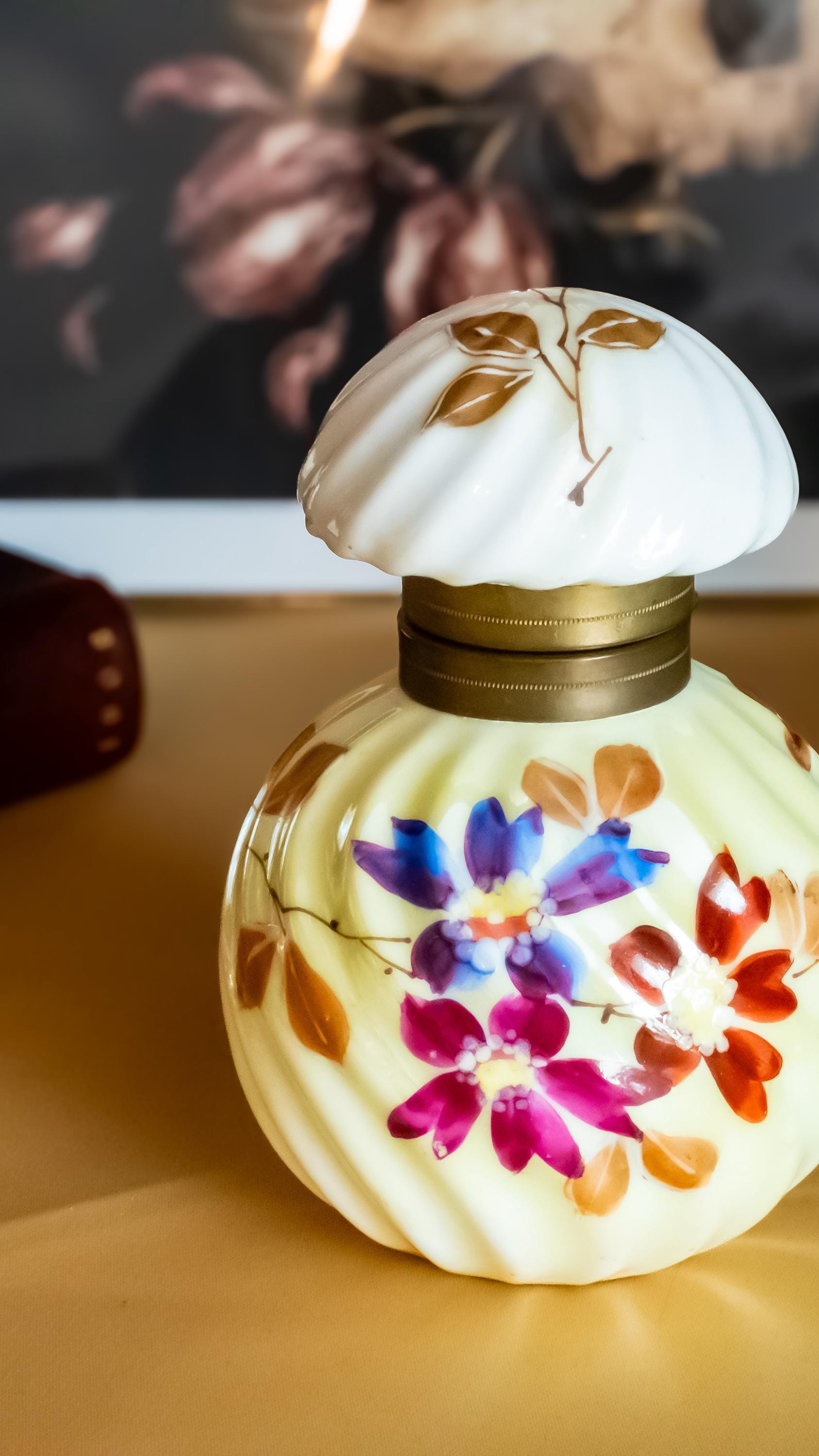 Antique pale yellow ribbed glass inkwell with hand-painted Victorian floral motifs in purple and red. The desk decor features a brass collar and a swirled white glass stopper, sitting on a writer's desk.