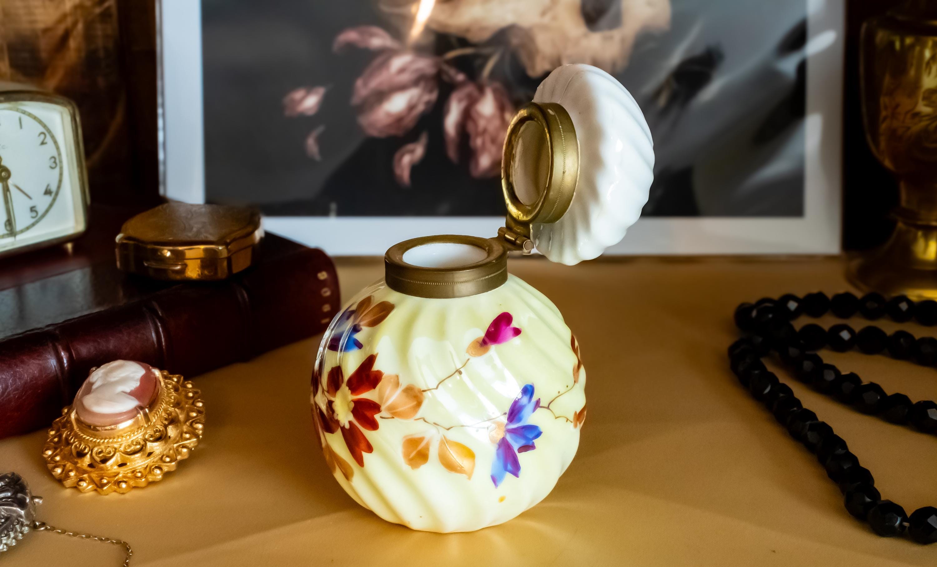 Antique pale yellow ribbed glass inkwell with hand-painted Victorian floral motifs in purple and red. The desk decor features a brass collar and a swirled white glass stopper, sitting on a writer's desk.
