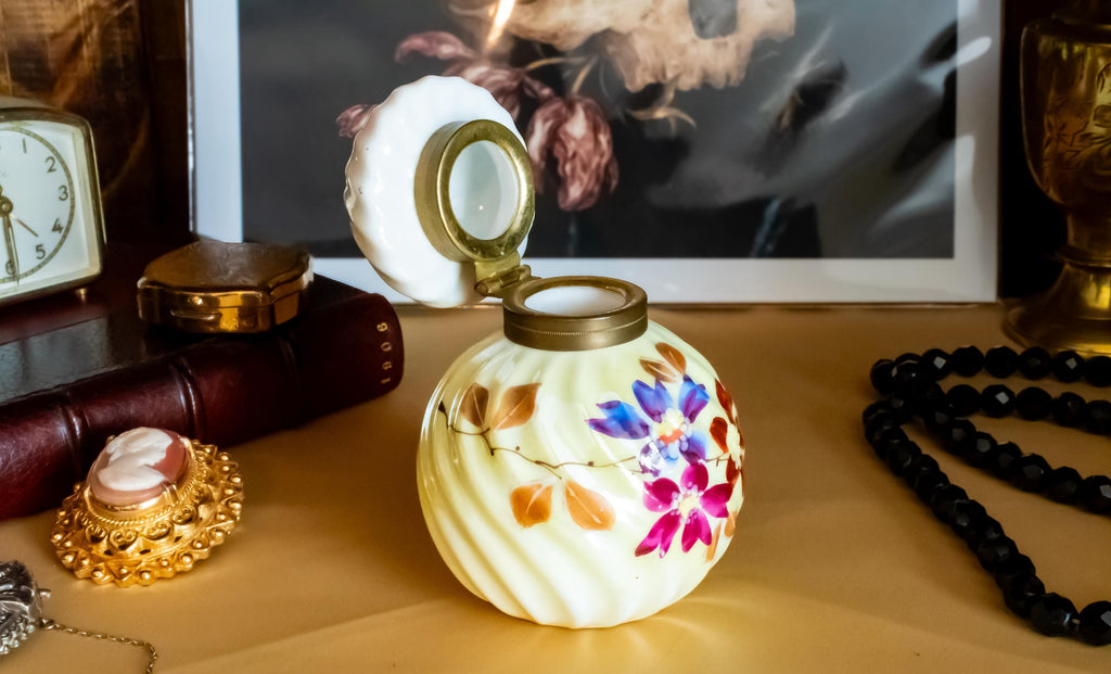 Antique pale yellow ribbed glass inkwell with hand-painted Victorian floral motifs in purple and red. The desk decor features a brass collar and a swirled white glass stopper, sitting on a writer's desk.