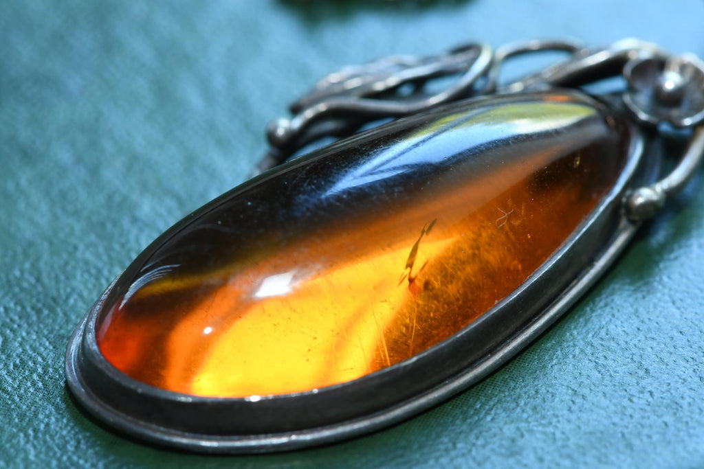 Large teardrop genuine Baltic amber pendant set in an oxidized sterling silver botanical leaf design. The brown witchy gothic necklace rests on a vintage green book alongside dark academia antiquities.