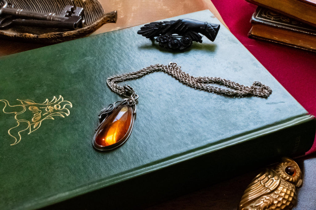 Large teardrop genuine Baltic amber pendant set in an oxidized sterling silver botanical leaf design. The brown witchy gothic necklace rests on a vintage green book alongside dark academia antiquities.