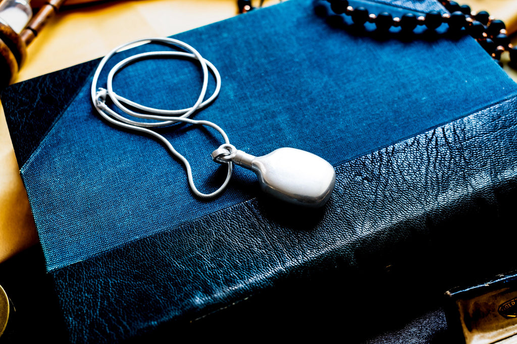 Heavy, smooth polished sterling silver antique potion flask pendant on a silver chain. The interactive Victorian scent bottle features a threaded cap and rests on a dark blue vintage book.