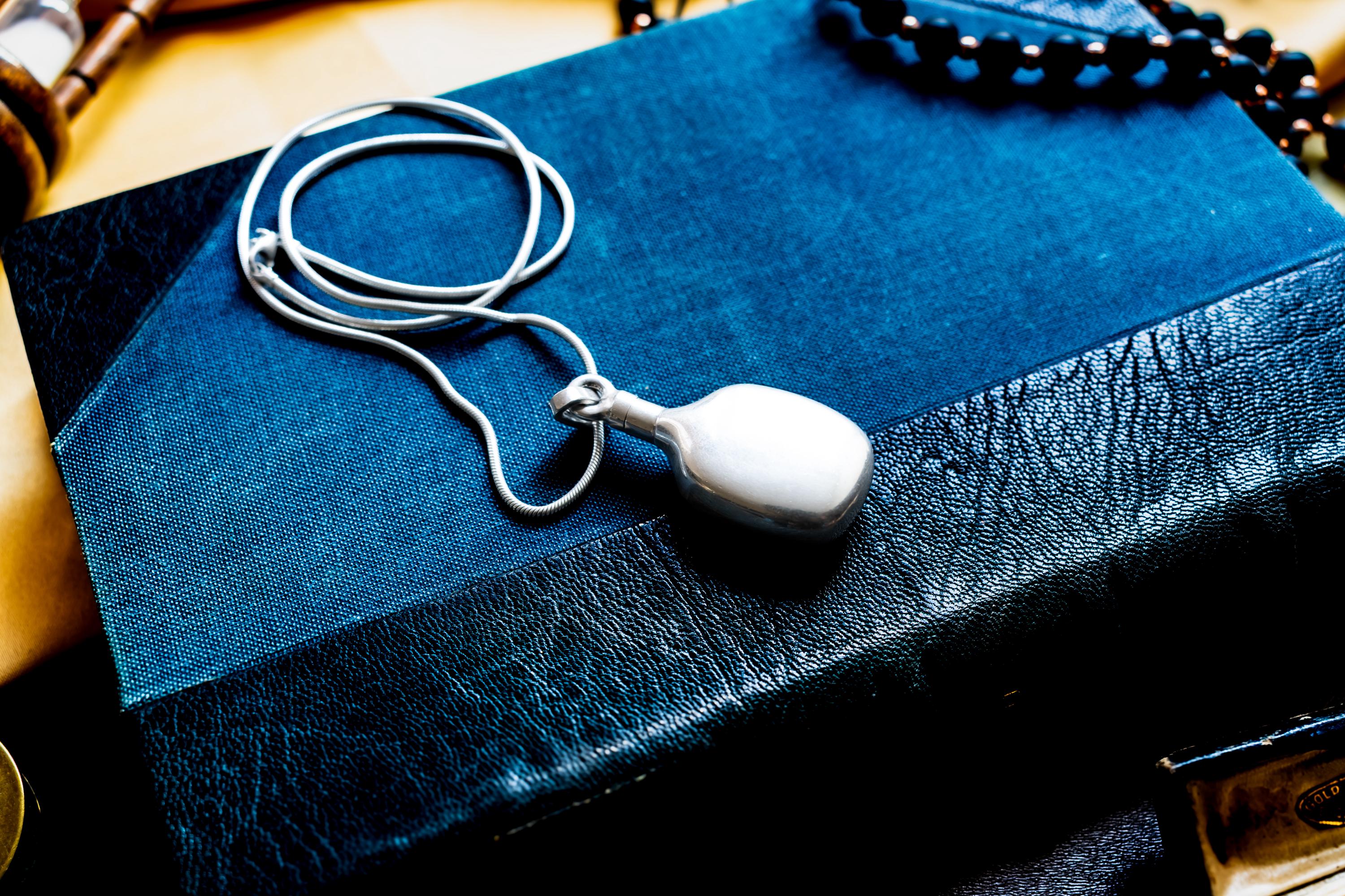 Heavy, smooth polished sterling silver antique potion flask pendant on a silver chain. The interactive Victorian scent bottle features a threaded cap and rests on a dark blue vintage book.