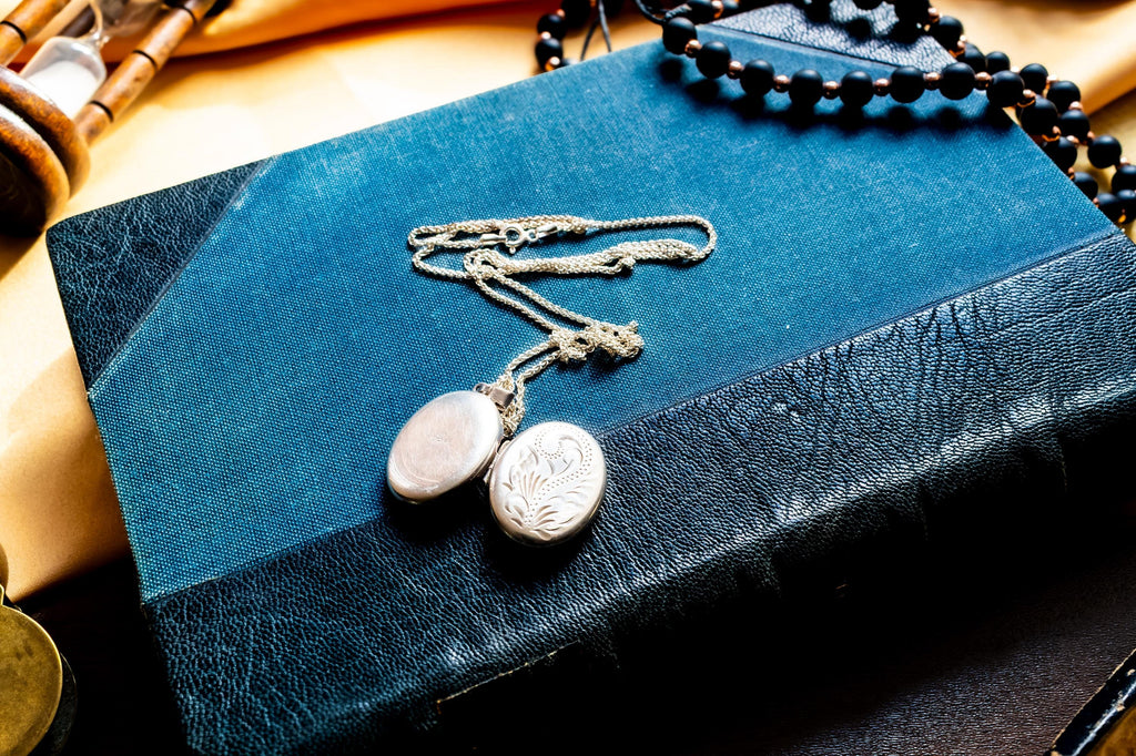 the image of the front and back of an oval silver locket pendant featuring an intricate engraved floral pattern on its front surface. The metal has a polished, reflective finish with deep etched lines creating a swirling leaf design. A small metal loop is attached at the top.