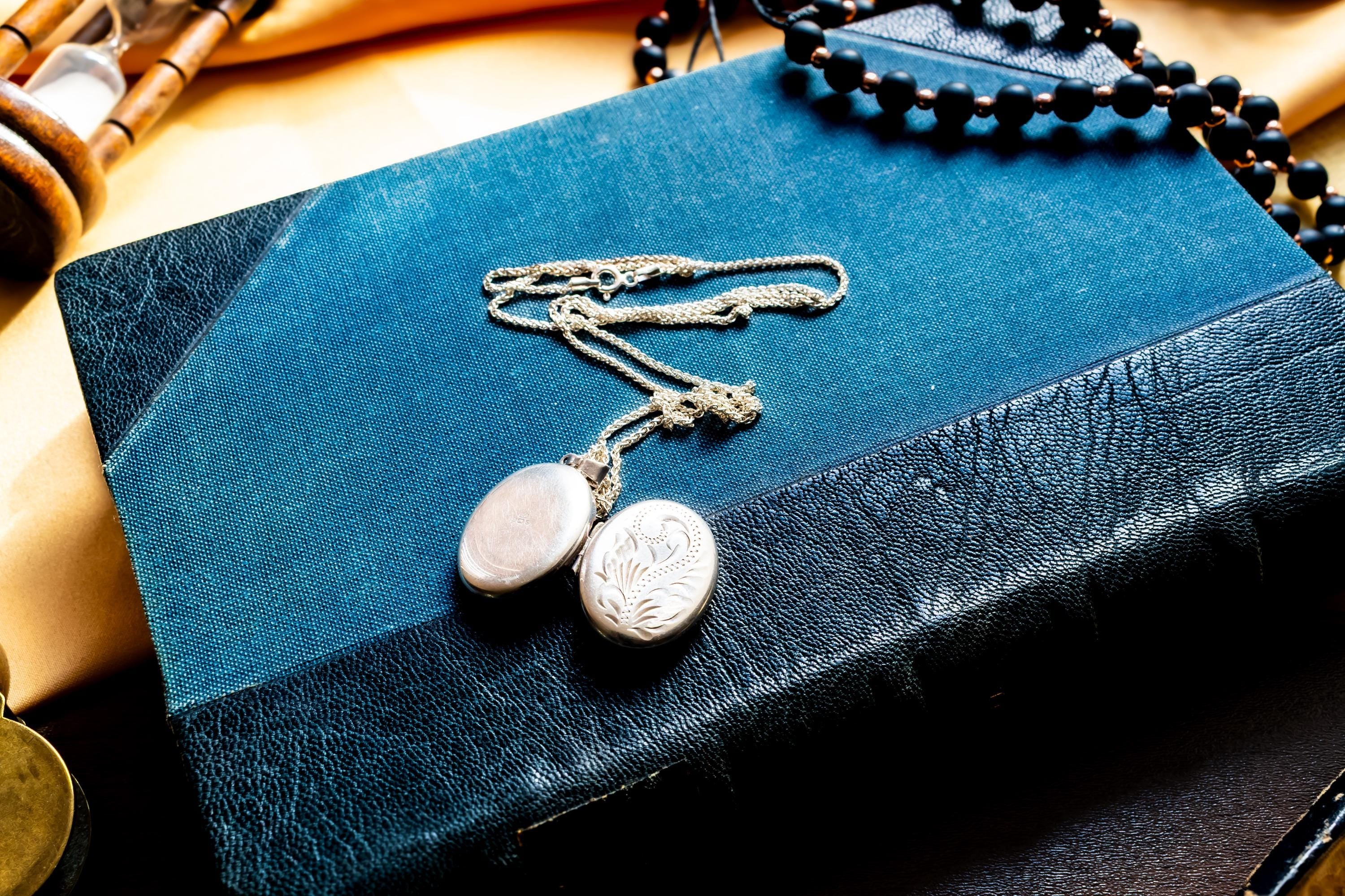 the image of the front and back of an oval silver locket pendant featuring an intricate engraved floral pattern on its front surface. The metal has a polished, reflective finish with deep etched lines creating a swirling leaf design. A small metal loop is attached at the top.