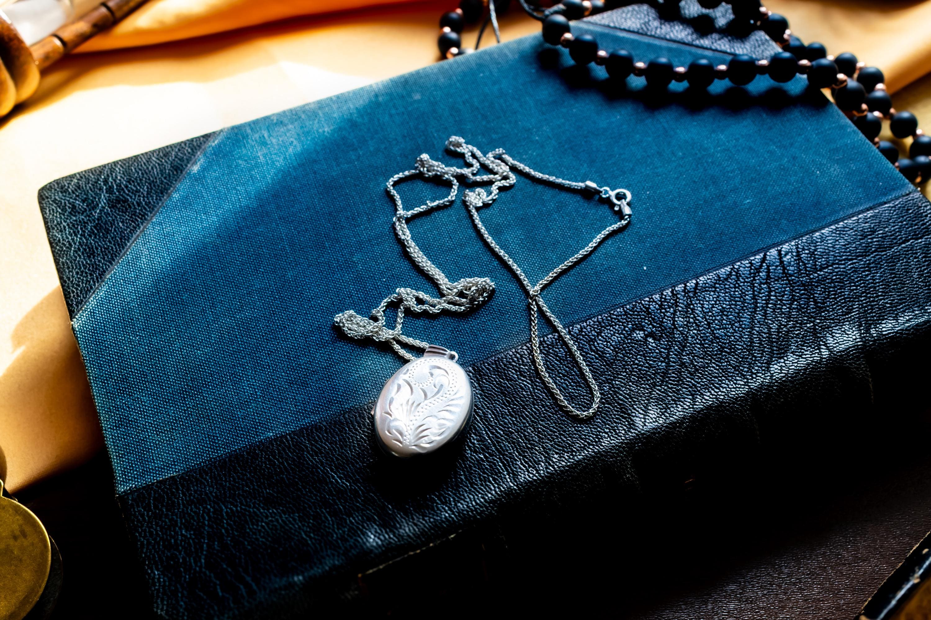 May include: An oval silver locket pendant featuring an intricate engraved floral pattern on its front surface. The metal has a polished, reflective finish with deep etched lines creating a swirling leaf design. A small metal loop is attached at the top.