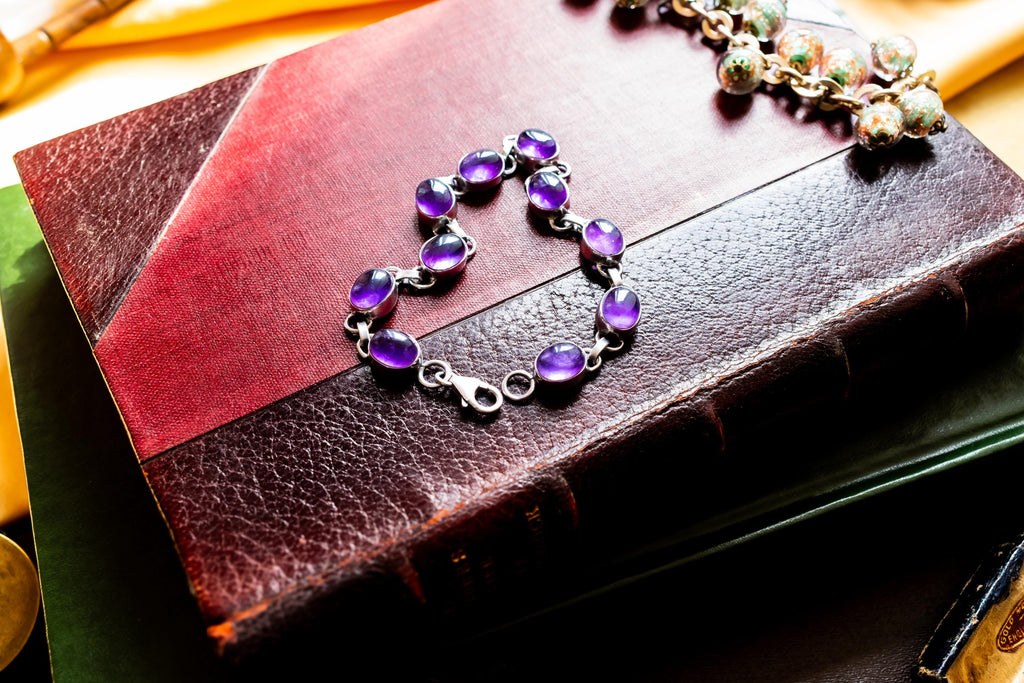 A silver bracelet featuring oval-shaped, deep purple amethyst gemstones set in polished metal links. The bracelet is draped on a red leather bound vintage book.