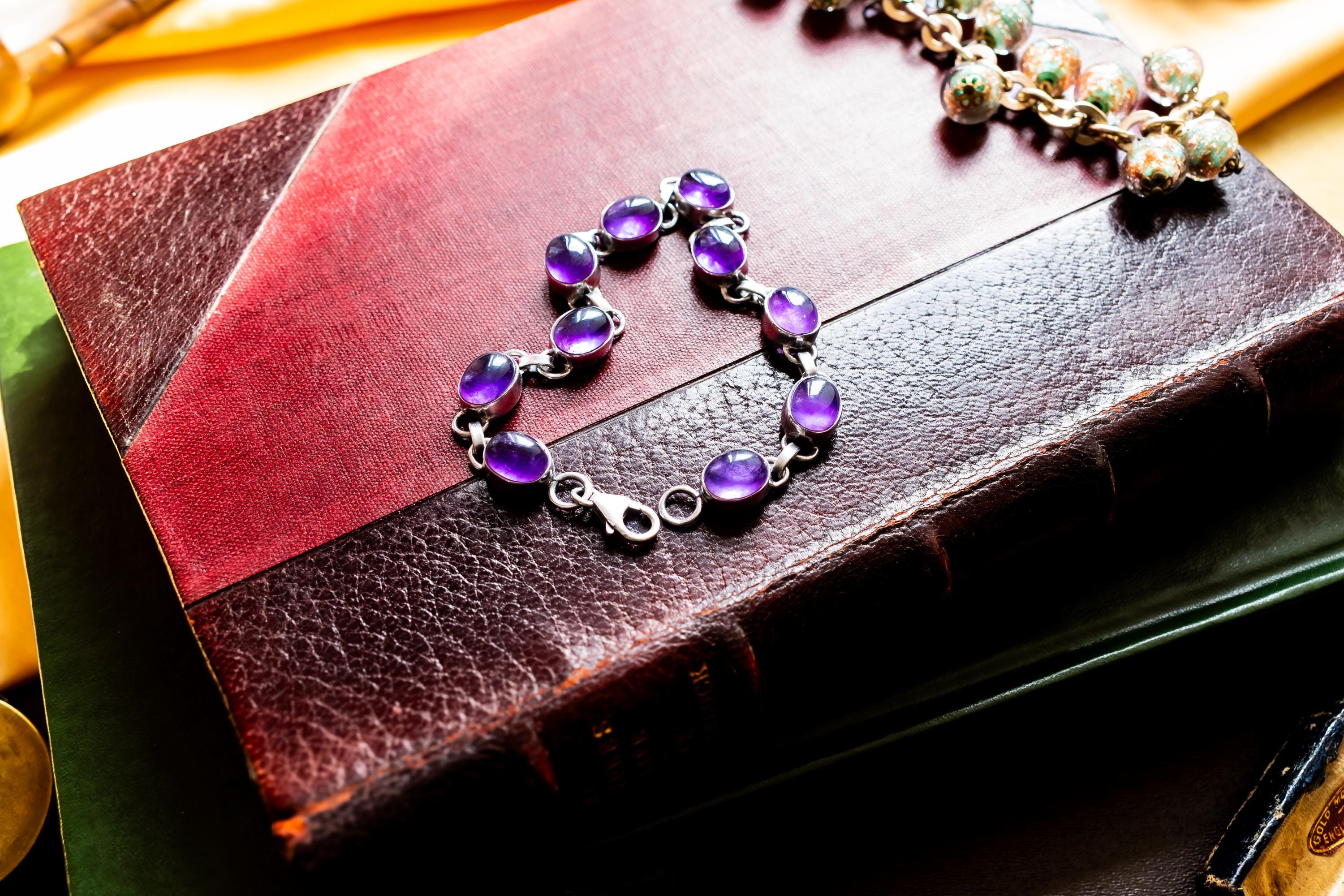 A silver bracelet featuring oval-shaped, deep purple amethyst gemstones set in polished metal links. The bracelet is draped on a red leather bound vintage book.