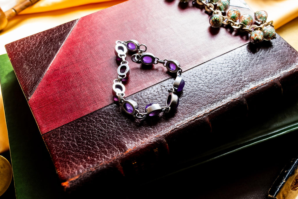 A silver bracelet featuring oval-shaped, deep purple amethyst gemstones set in polished metal links. The bracelet is draped on a red leather bound vintage book.