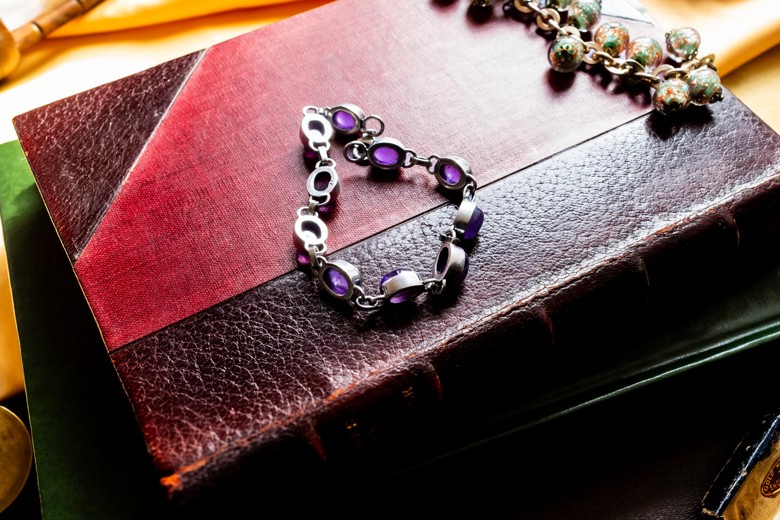 A silver bracelet featuring oval-shaped, deep purple amethyst gemstones set in polished metal links. The bracelet is draped on a red leather bound vintage book.