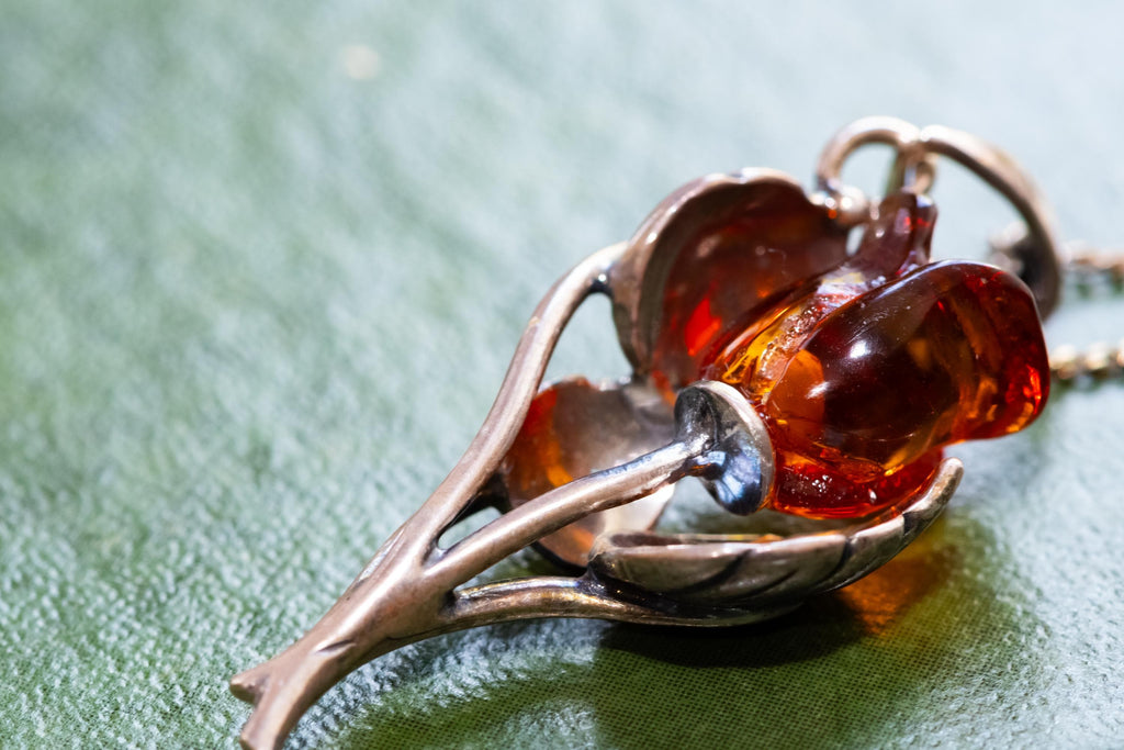 A vintage 925 sterling silver necklace featuring a carved Baltic amber rose pendant, beautifully staged resting on the cover of an antique green book with a dark carved brooch in the background.