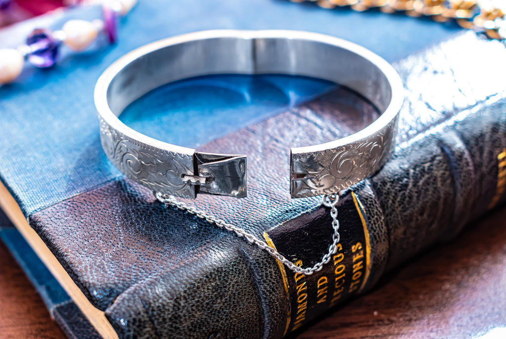 A vintage 1979 Sheffield sterling silver hinged cuff bracelet featuring intricate scrollwork engraving and a delicate safety chain, resting on the spine of an antique dark green book titled 'Diamonds and Precious Stones'.