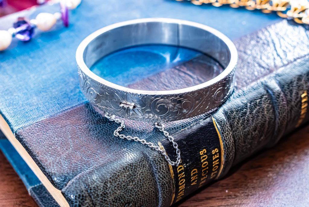 A vintage 1979 Sheffield sterling silver hinged cuff bracelet featuring intricate scrollwork engraving and a delicate safety chain, resting on the spine of an antique dark green book titled 'Diamonds and Precious Stones'.