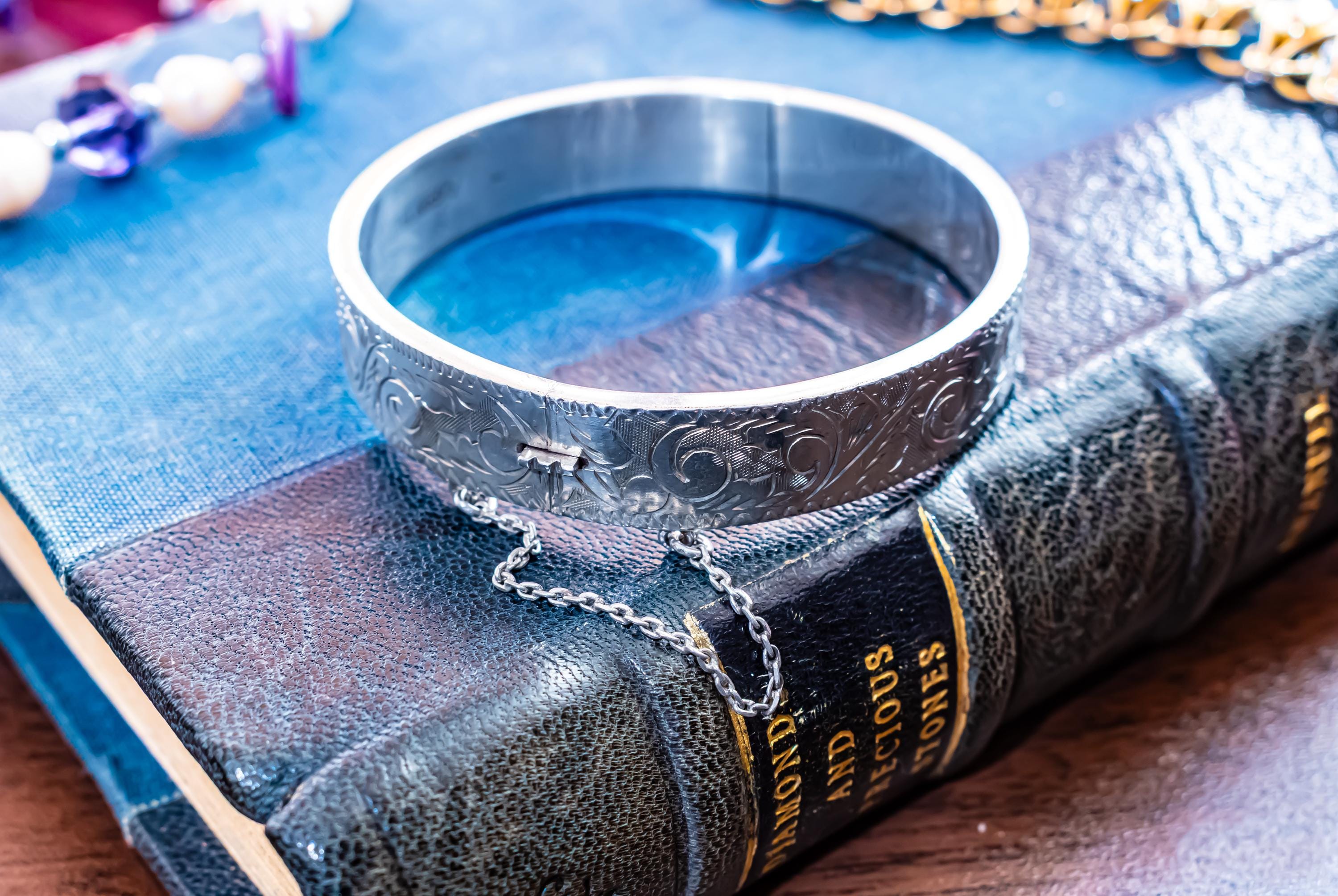 A vintage 1979 Sheffield sterling silver hinged cuff bracelet featuring intricate scrollwork engraving and a delicate safety chain, resting on the spine of an antique dark green book titled 'Diamonds and Precious Stones'.