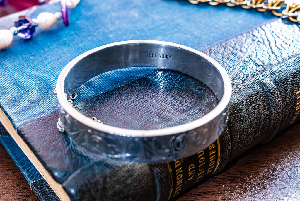 A vintage 1979 Sheffield sterling silver hinged cuff bracelet featuring intricate scrollwork engraving and a delicate safety chain, resting on the spine of an antique dark green book titled 'Diamonds and Precious Stones'.
