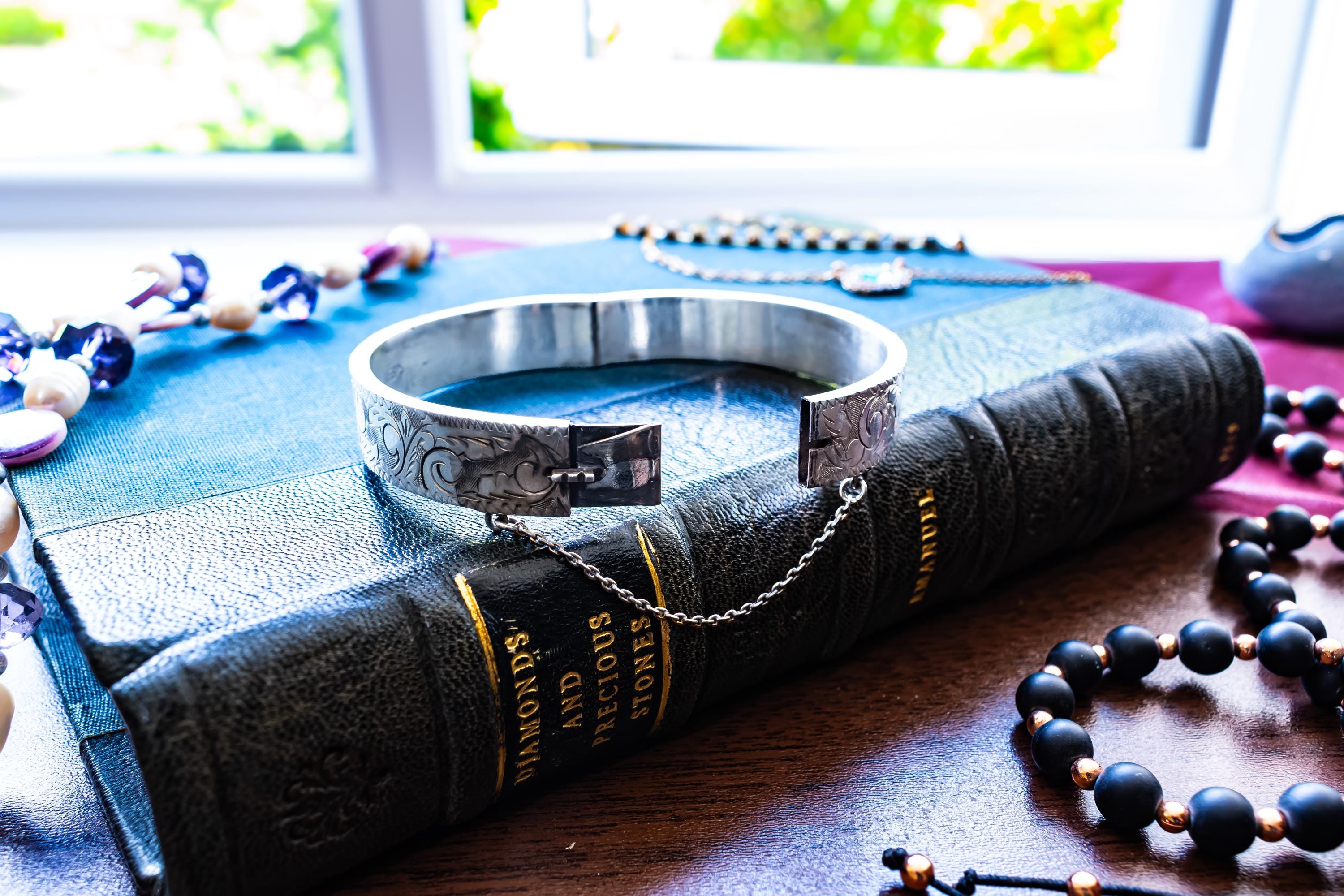 A vintage 1979 Sheffield sterling silver hinged cuff bracelet featuring intricate scrollwork engraving and a delicate safety chain, resting on the spine of an antique dark green book titled 'Diamonds and Precious Stones'.