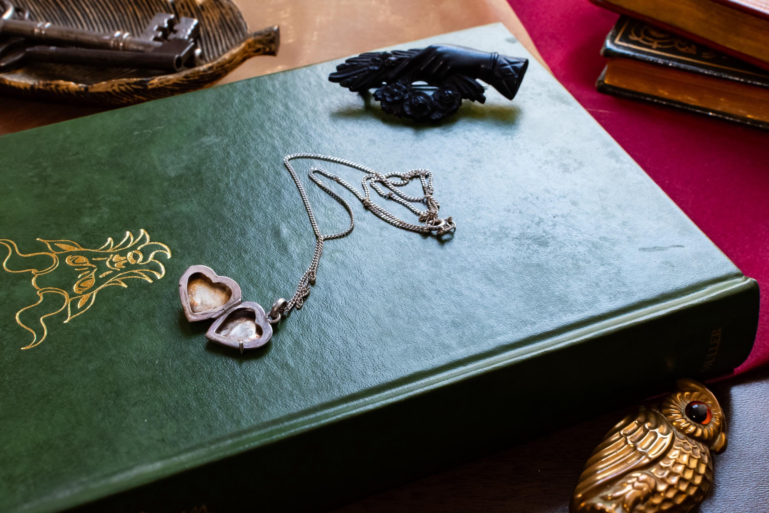A vintage silver heart locket necklace featuring a small embossed teddy bear design, resting beautifully on a green antique book with gold embossing. A small brass owl and a carved black hand brooch sit in the background.