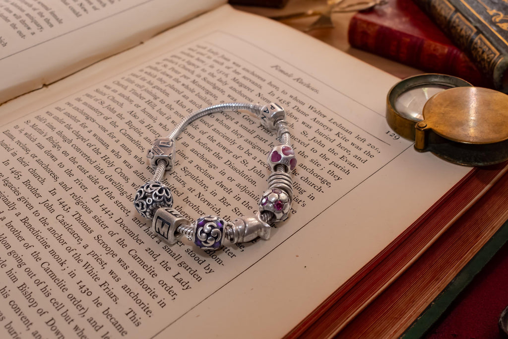 Authentic Pandora sterling silver snake chain bracelet loaded with nine various charms including a letter M and purple enamel beads, resting on an open antique book.