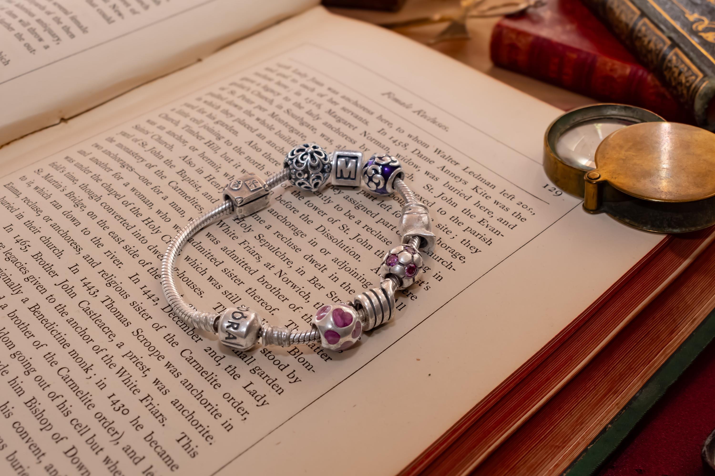 Authentic Pandora sterling silver snake chain bracelet loaded with nine various charms including a letter M and purple enamel beads, resting on an open antique book.