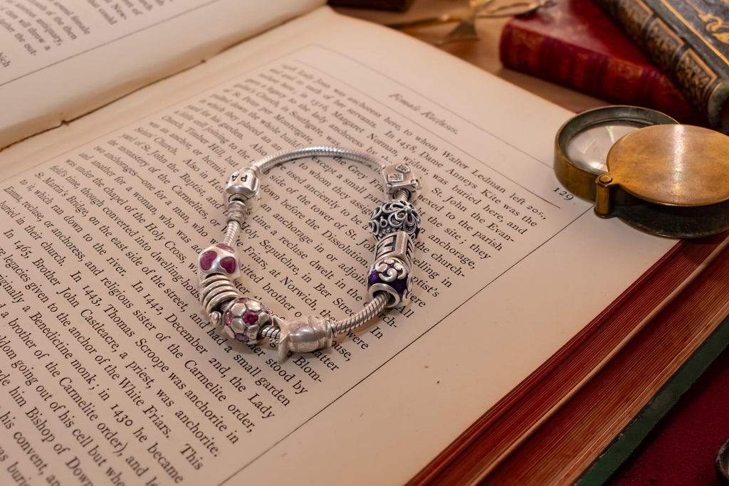 Authentic Pandora sterling silver snake chain bracelet loaded with nine various charms including a letter M and purple enamel beads, resting on an open antique book.