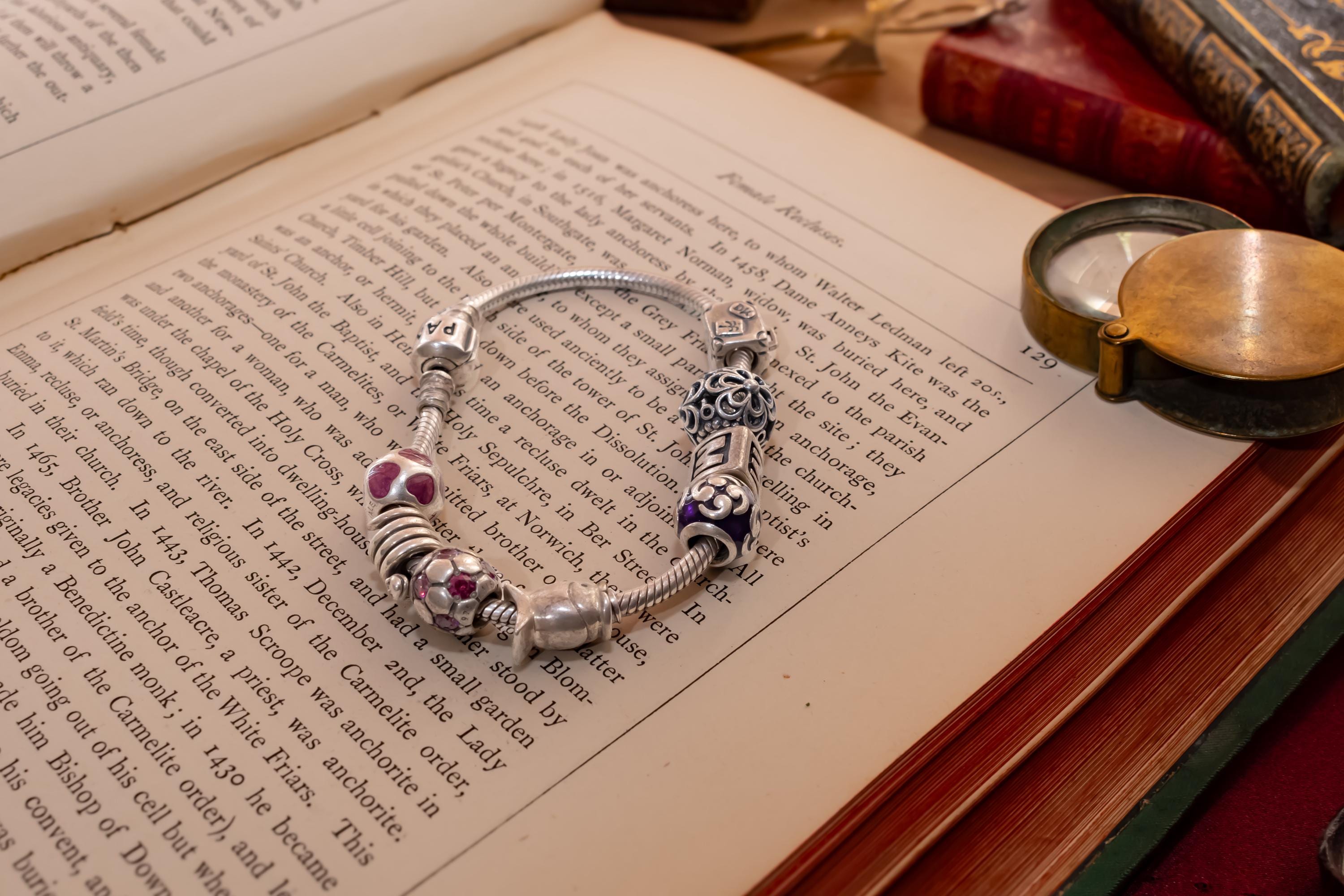 Authentic Pandora sterling silver snake chain bracelet loaded with nine various charms including a letter M and purple enamel beads, resting on an open antique book.