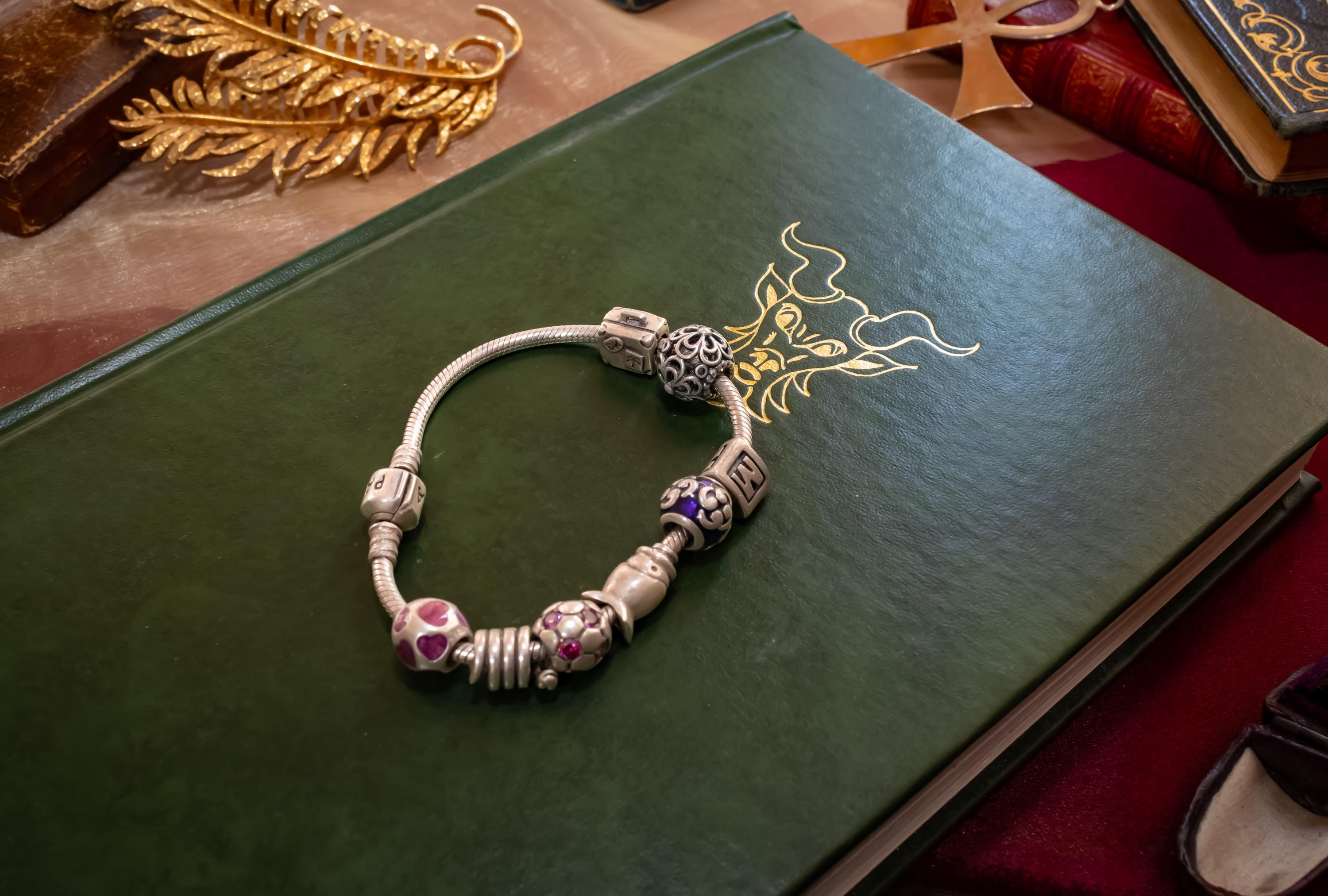 Authentic Pandora sterling silver snake chain bracelet loaded with nine various charms including a letter M and purple enamel beads, resting on an open antique book.