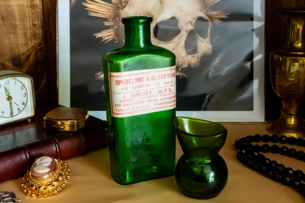 Two vintage green glass apothecary bottles, one tall with a vintage label and one small, sitting on a desk beside a brass clock and skull art.