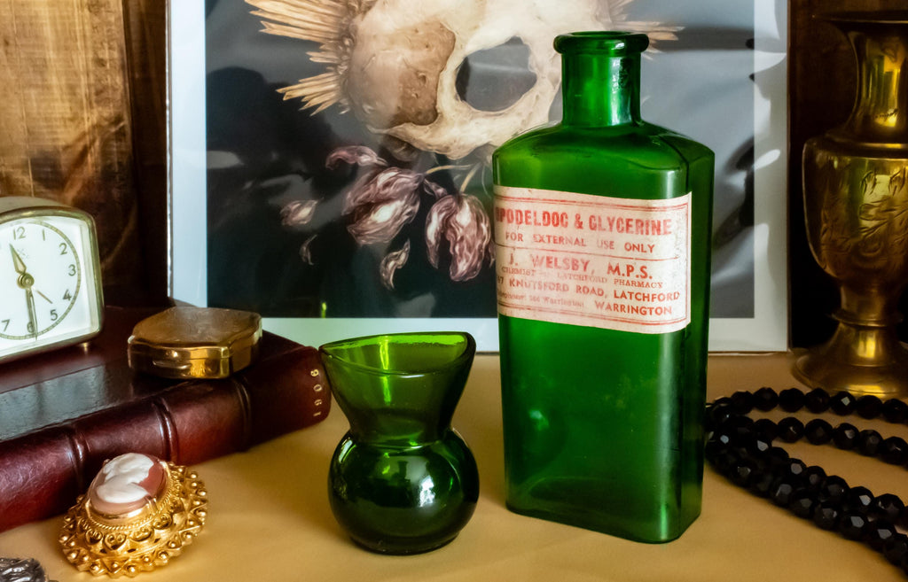 Two vintage green glass apothecary bottles, one tall with a vintage label and one small, sitting on a desk beside a brass clock and skull art.