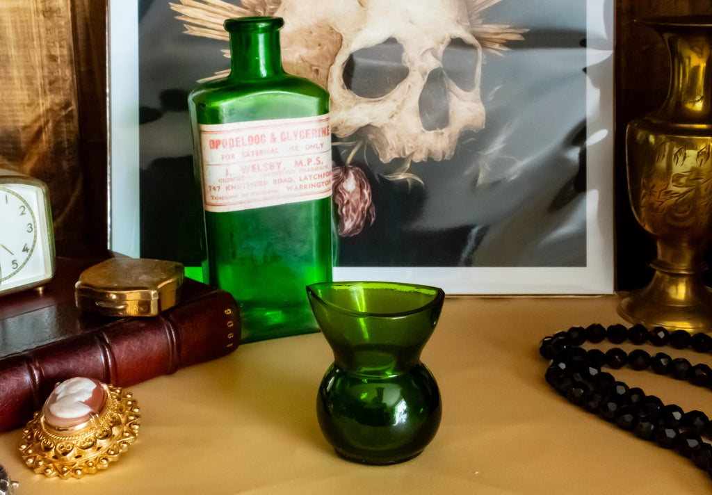 Two vintage green glass apothecary bottles, one tall with a vintage label and one small, sitting on a desk beside a brass clock and skull art.