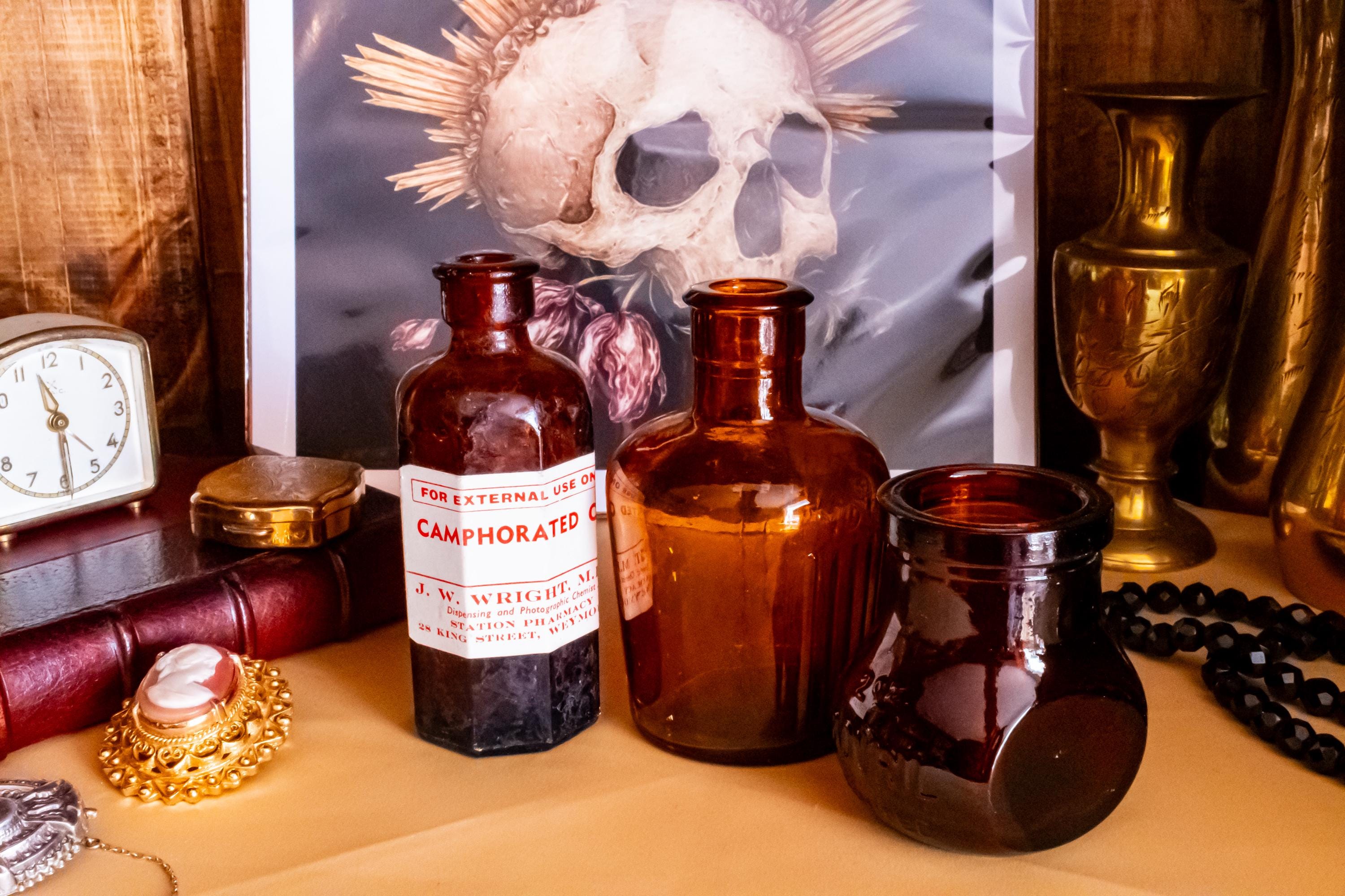 Three vintage amber glass apothecary bottles in varying shapes and sizes, one retaining a vintage label, displayed near a brass vase and skull artwork.